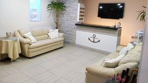 Uma sala de estar moderna com um sofá bege, uma mesinha com abajur, uma planta em vaso e uma TV fixada na parede.