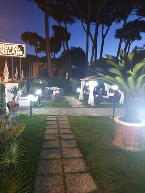 Entrada de un hotel con un camino empedrado que lleva a una zona de estar, rodeada de vegetación y árboles.
