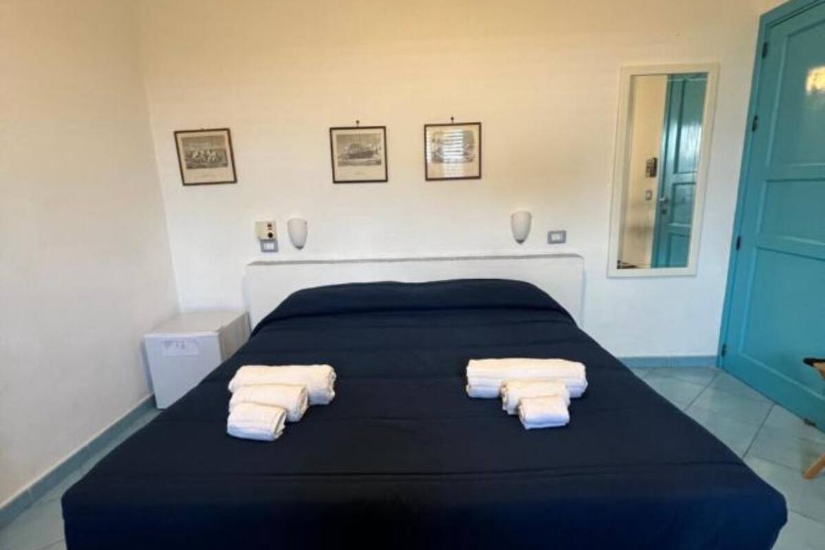 A double bed with a black bedspread and two towels, a small bedside table with a lamp, and three framed pictures on the wall.