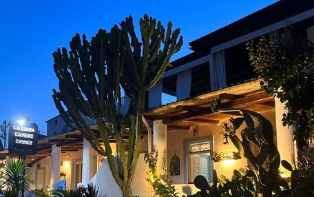 A hotel with a large cactus in the foreground, a sign reading "LA GIARA CAMERE ZIMMER," and a well-lit facade at dusk.