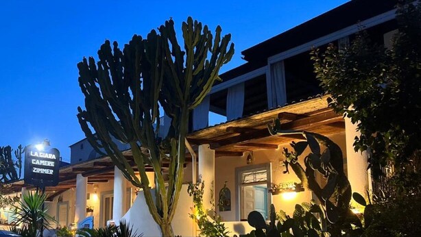 A hotel with a large cactus in the foreground, a sign reading "LA GIARA CAMERE ZIMMER," and a well-lit facade at dusk. - La Giara Vulcano (Lipari)