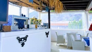 A covered outdoor seating area with a thatched roof, white wicker furniture, and a bar area with a blue and white tiled backsplash. - La Giara Vulcano (Lipari)