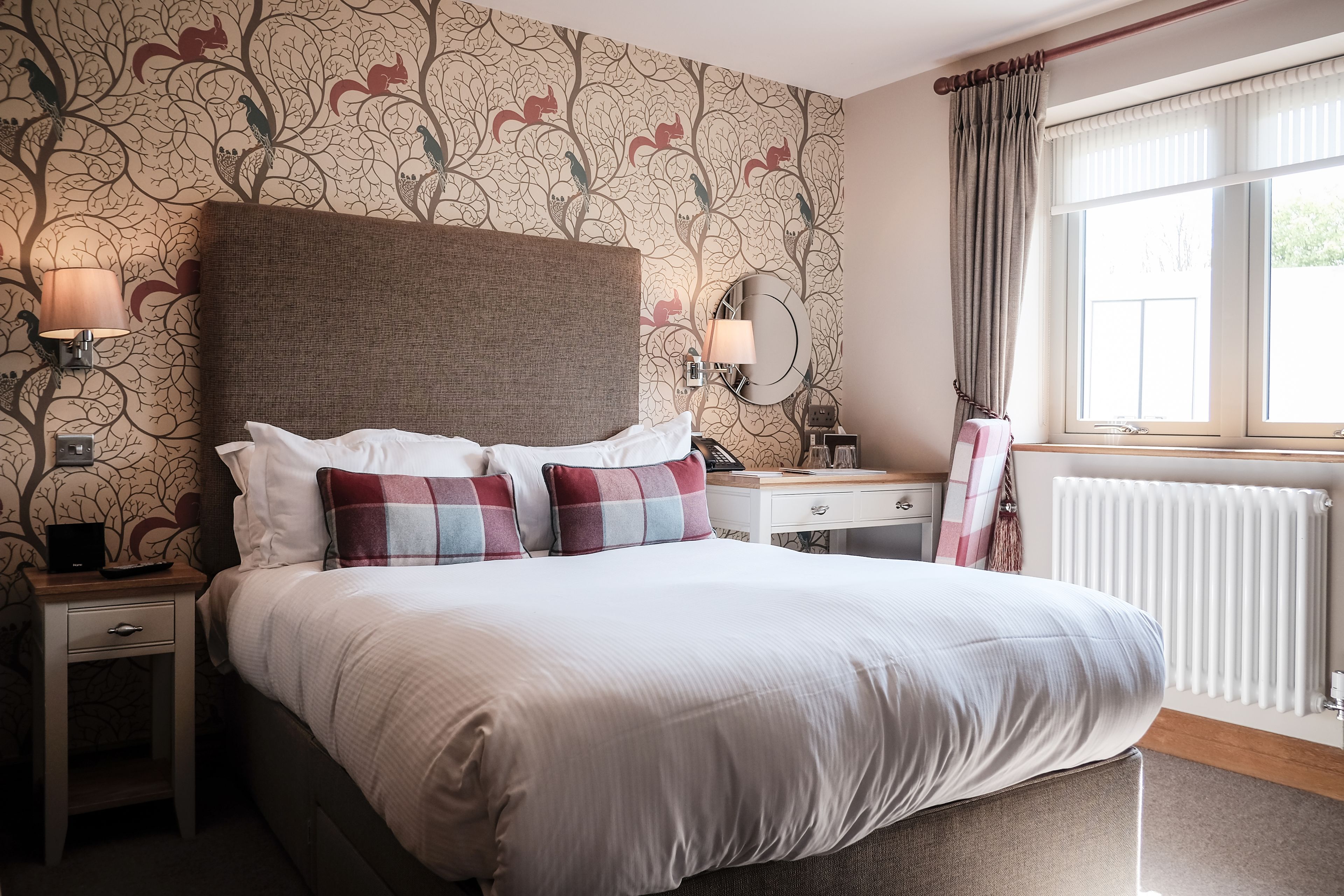 A neatly made bed with white and plaid pillows, a wooden nightstand with a lamp, a dresser with a mirror, and a window with curtains.
