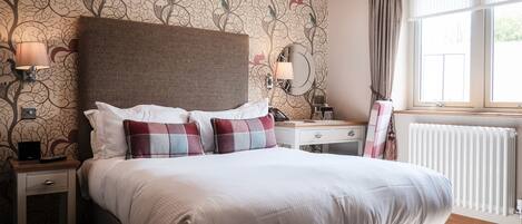 A neatly made bed with white and plaid pillows, a wooden nightstand with a lamp, a dresser with a mirror, and a window with curtains.
