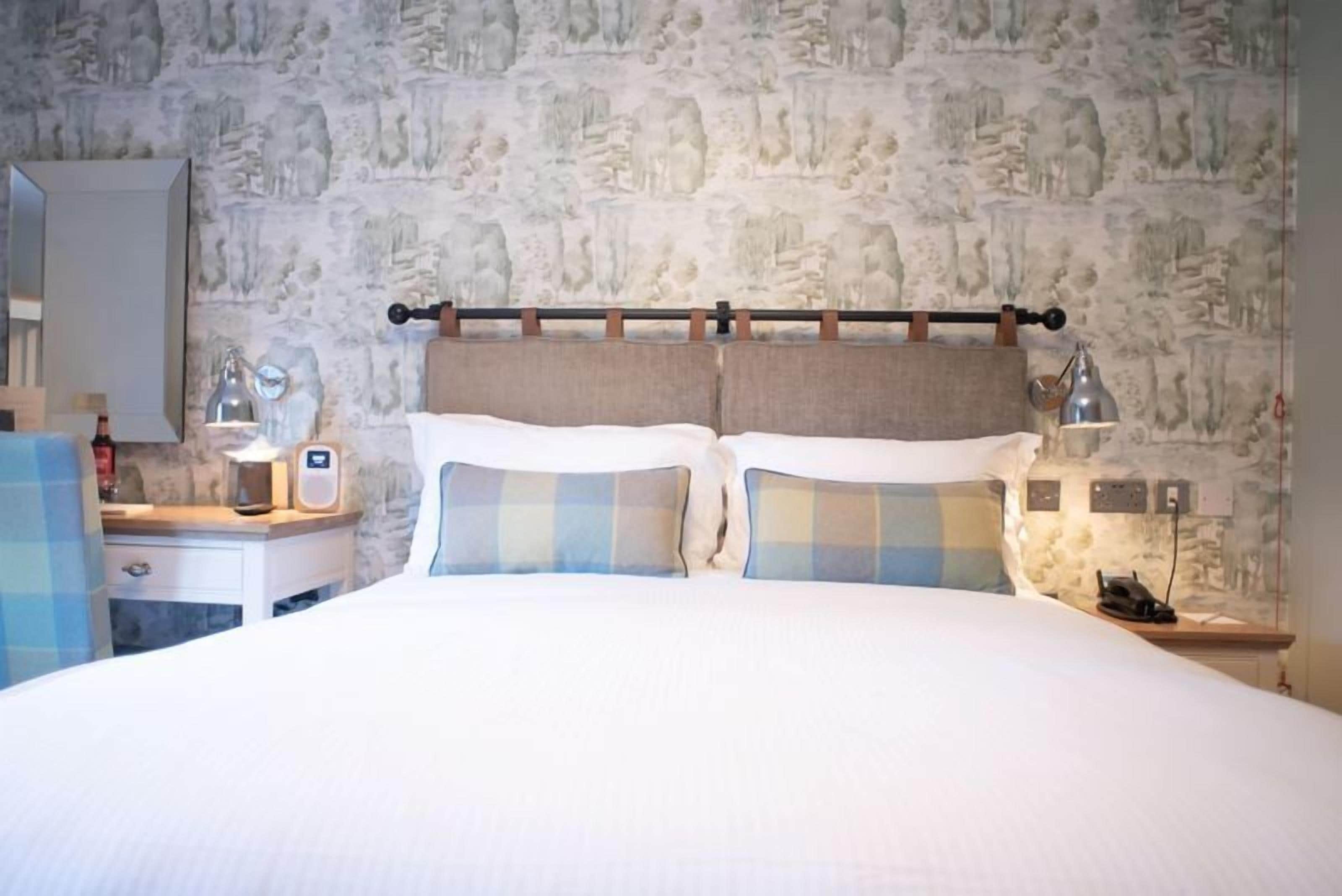 Una habitación de hotel con pared de piedra, una cama con almohadones a cuadros, una mesita de noche con lámpara y un espejo.