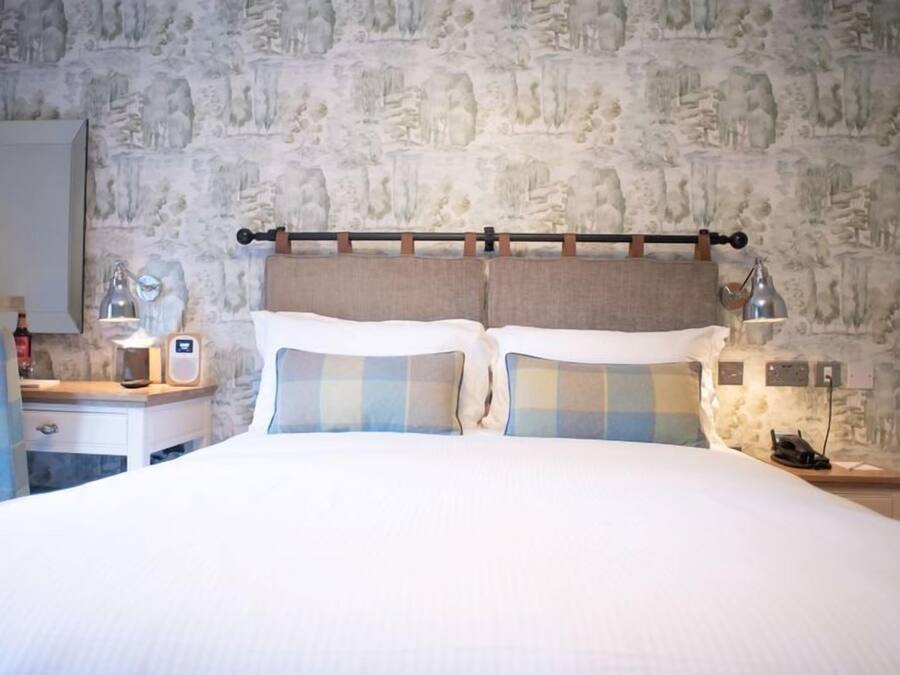 Una habitación de hotel con pared de piedra, una cama con almohadones a cuadros, una mesita de noche con lámpara y un espejo.