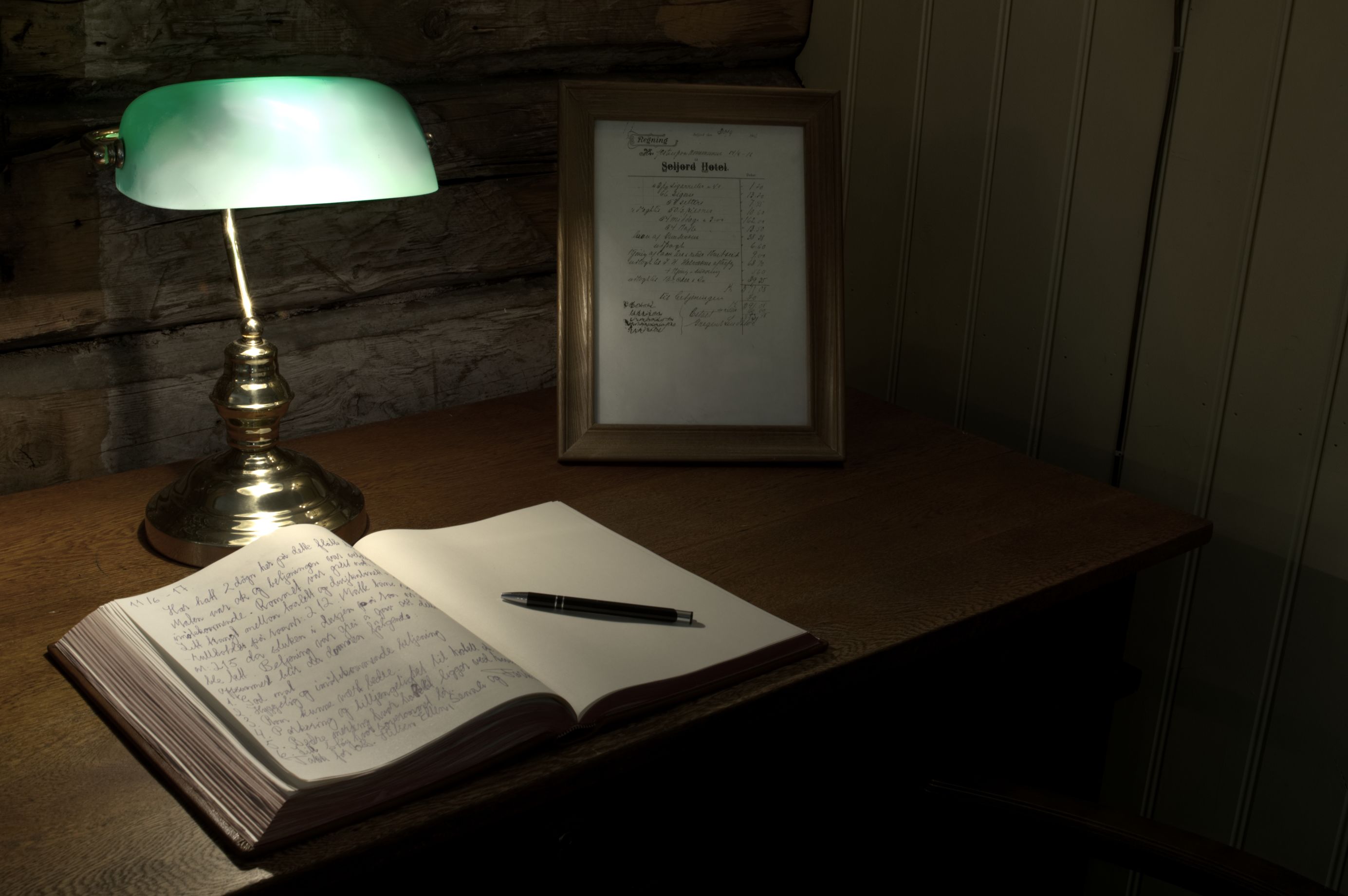 Una mesa de madera con un menú enmarcado, una lámpara de escritorio iluminada en verde y un cuaderno abierto con un bolígrafo encima.
