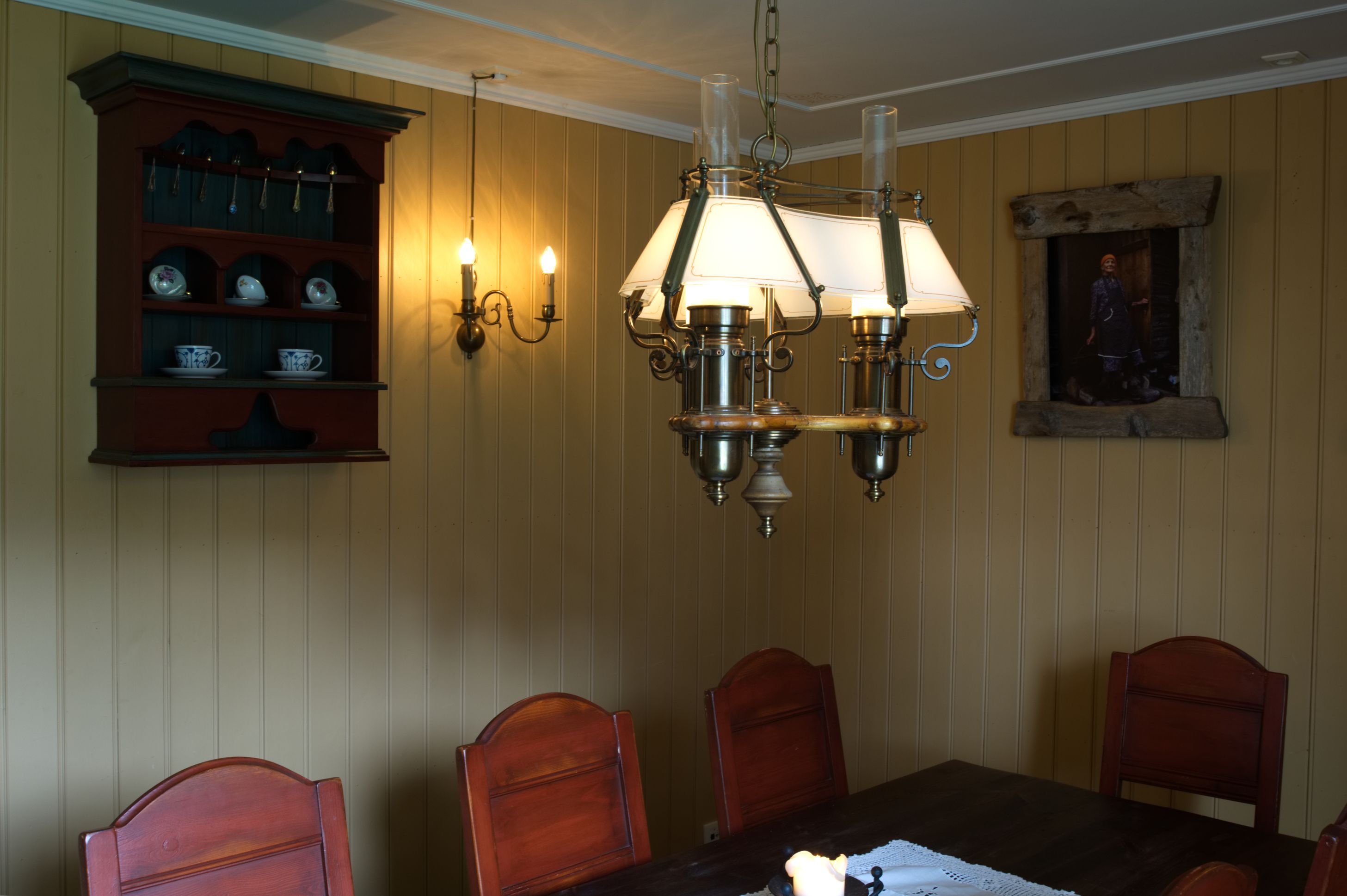 Une salle à manger avec une table en bois, quatre chaises, un lustre, un meuble mural contenant de la vaisselle et un tableau encadré.