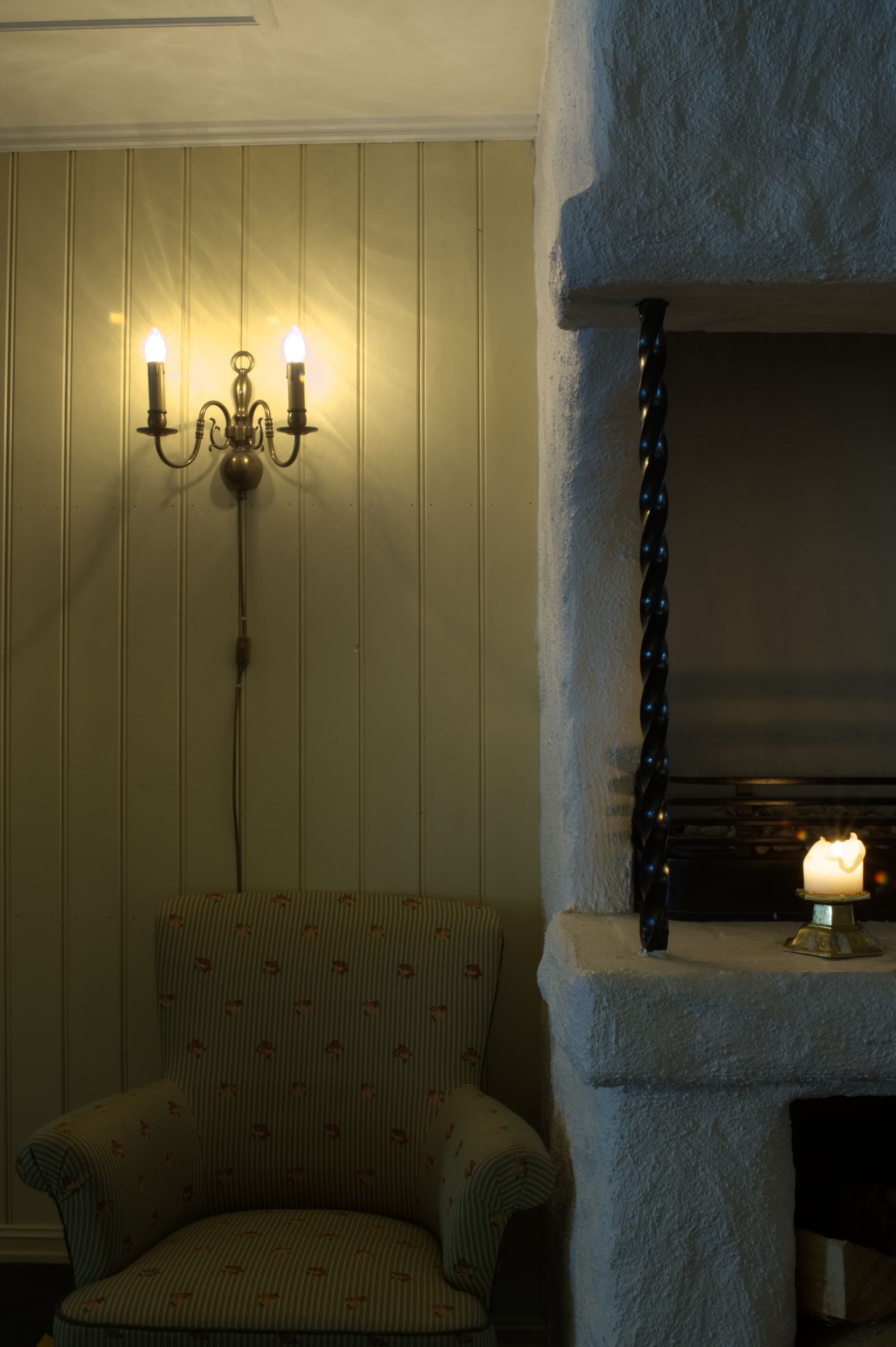 A room with a patterned armchair, a wall-mounted chandelier, and a stone fireplace with a lit candle.
