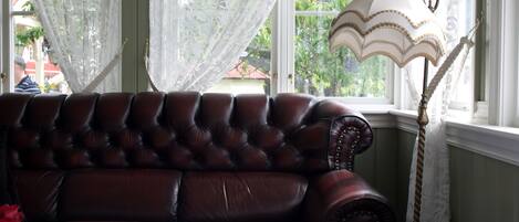 A dark brown leather sofa with a tufted design, positioned by a window with sheer white curtains. A table lamp with a white shade and gold trim is placed beside the sofa.