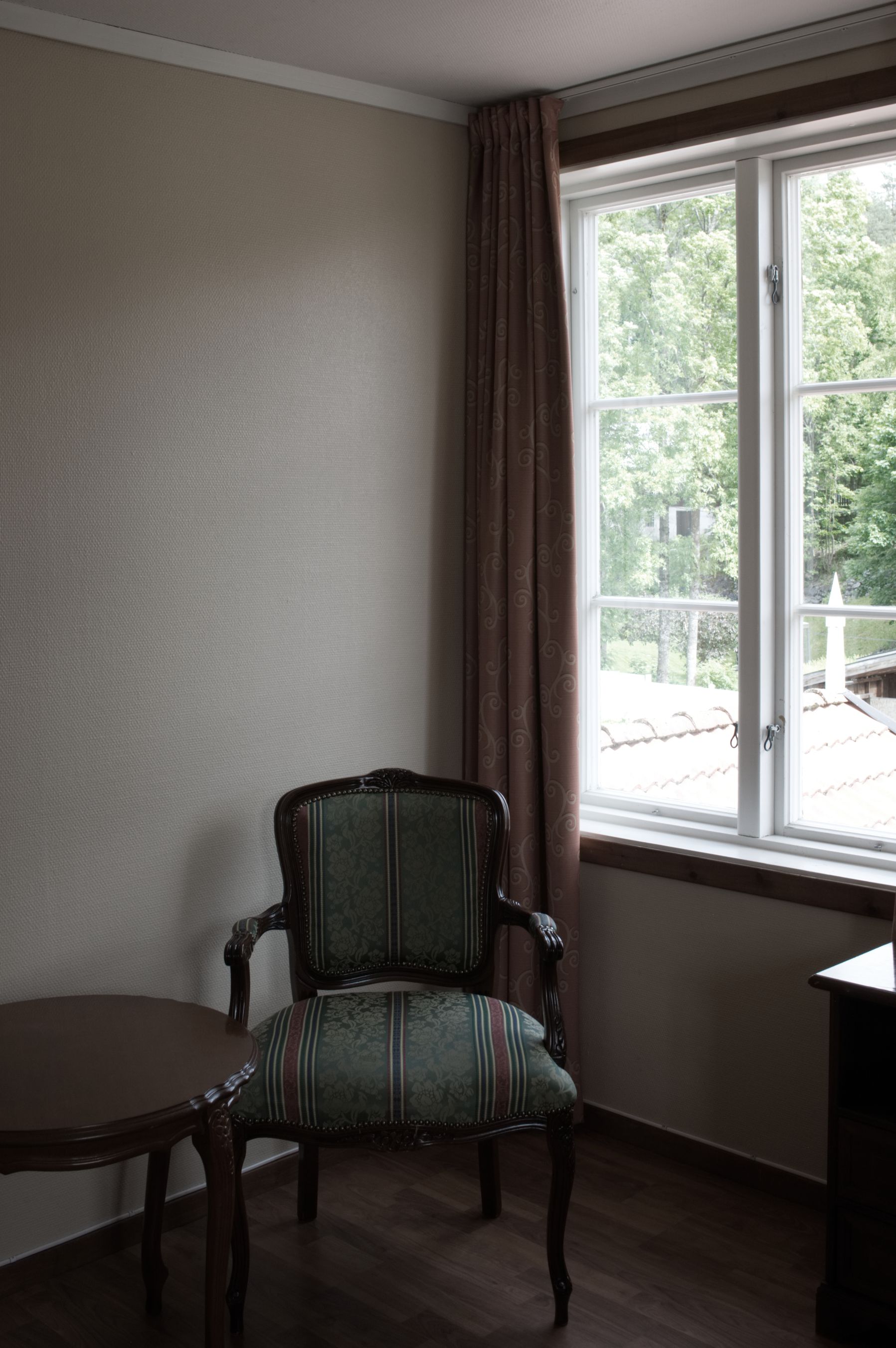 Una habitación de esquina con una silla de madera, una mesita y una ventana con cortinas.