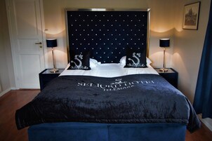 A hotel room with a large bed, a dark headboard with a pattern, two bedside tables with lamps, and a framed picture on the wall.
