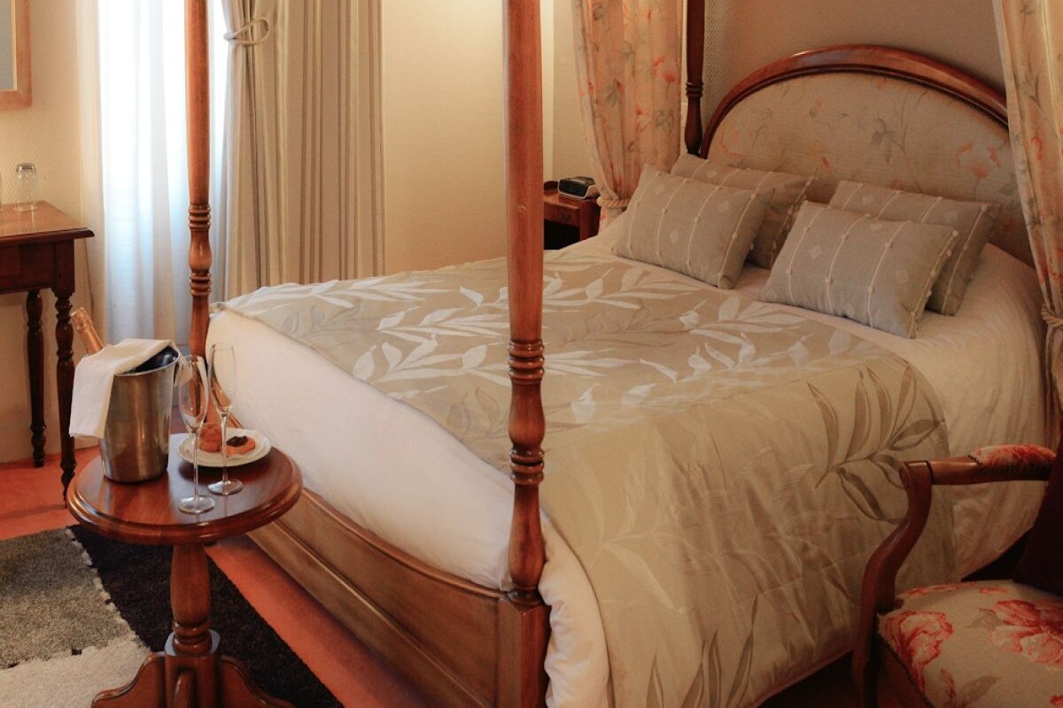 A four-poster bed with a floral bedspread, a bedside table with a bottle and glasses, and a nightstand with a lamp.