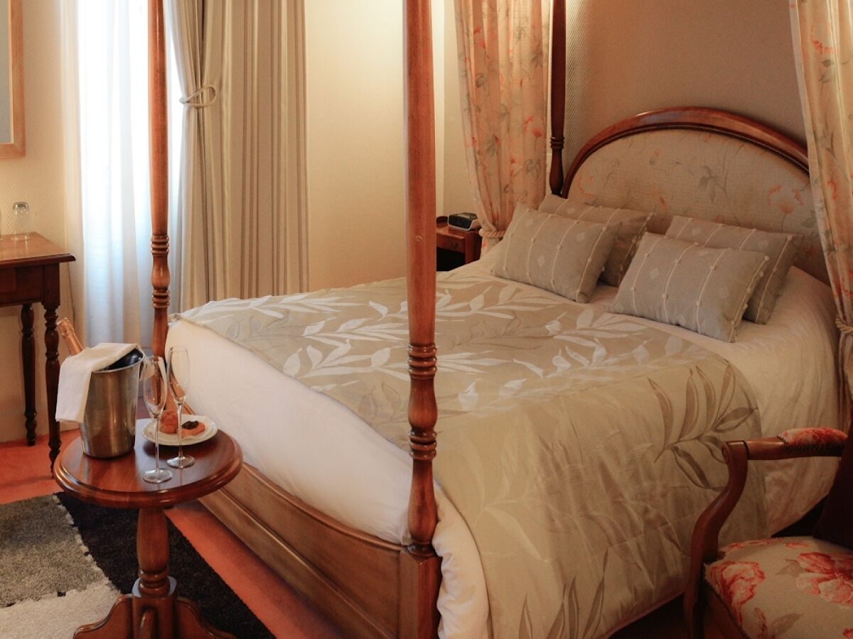 A four-poster bed with a floral bedspread, a bedside table with a bottle and glasses, and a nightstand with a lamp.