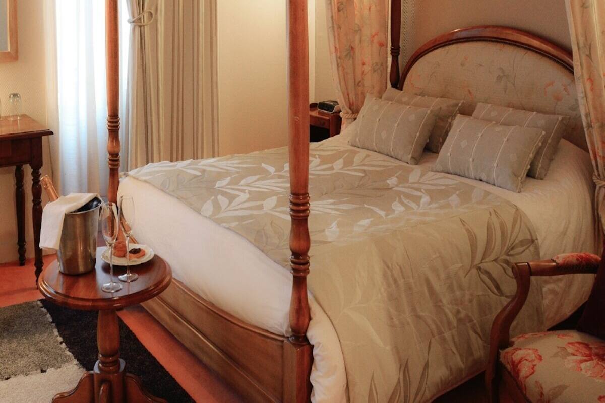 A four-poster bed with a floral bedspread, a bedside table with a bottle and glasses, and a nightstand with a lamp.
