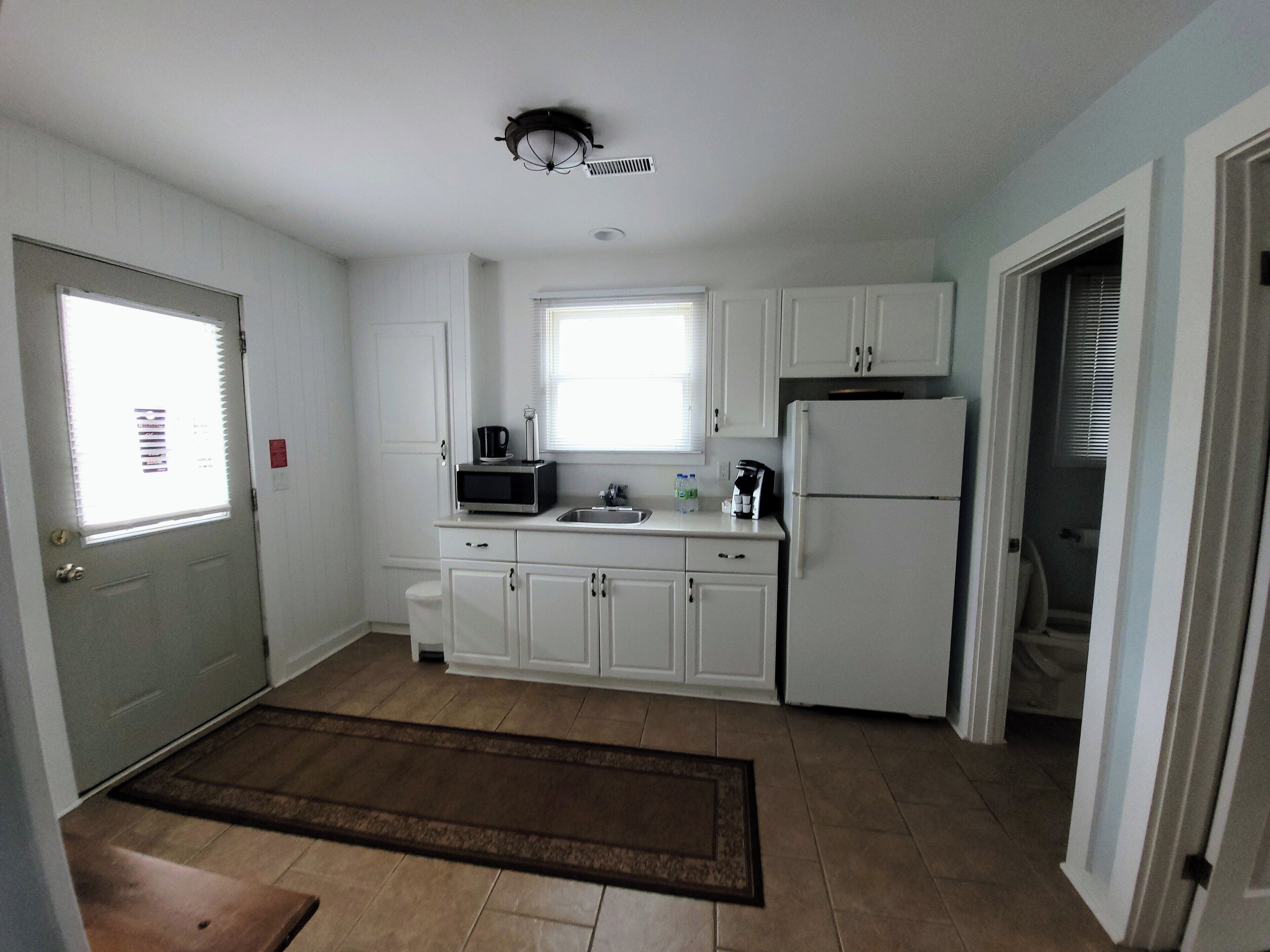 Cocina compacta con armarios blancos, microondas, refrigerador y ventana con persianas.