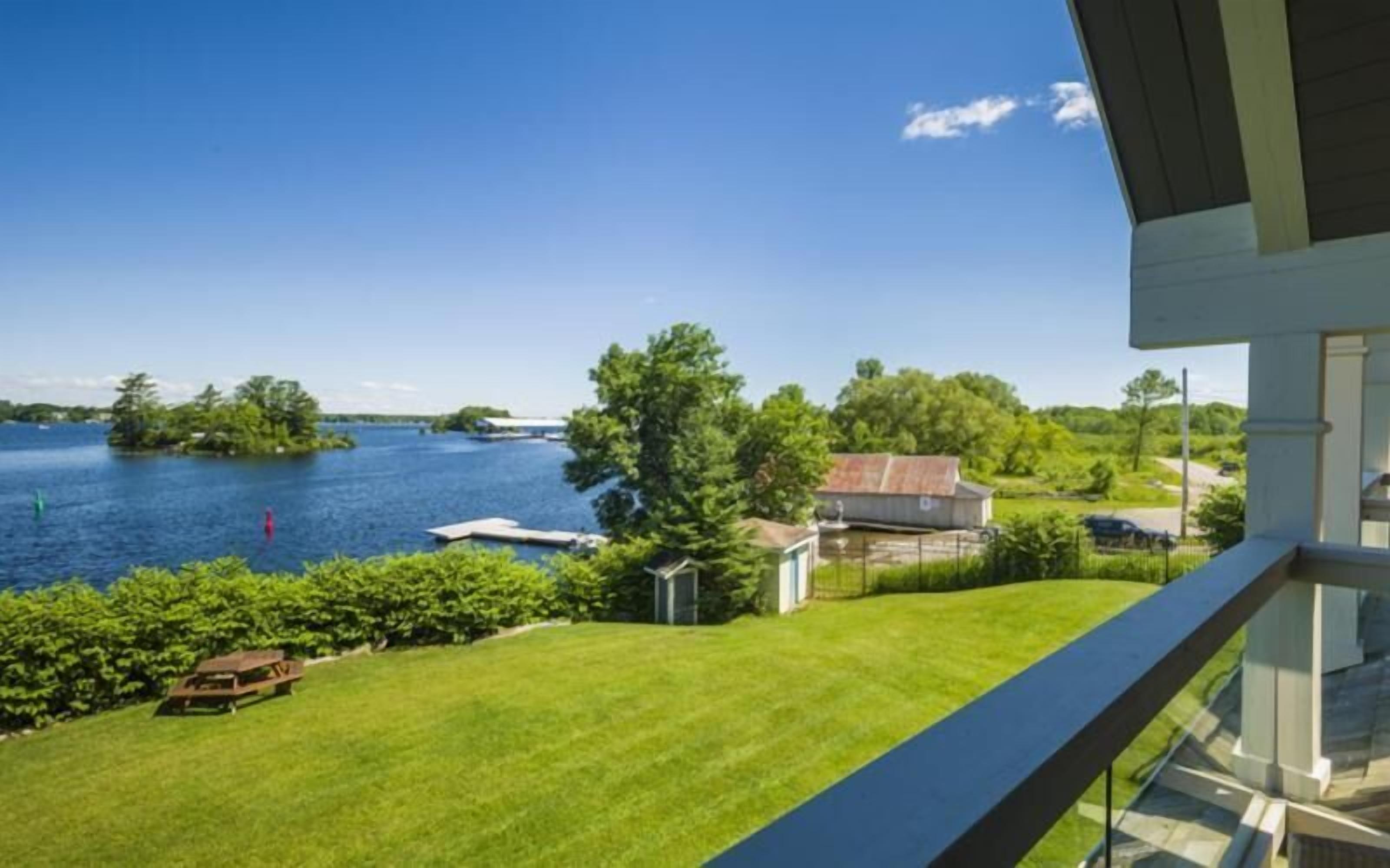 En utsikt från en balkong med utsikt över en sjö, med en gräsyta, en liten brygga och ett hus i bakgrunden.