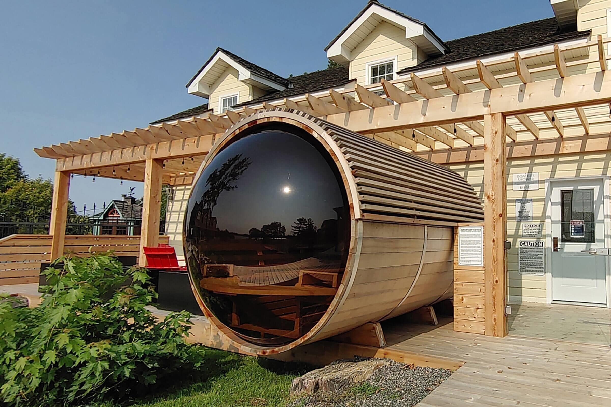 A large cylindrical structure with a reflective surface, a wooden pergola, and a building with a sign in the background.