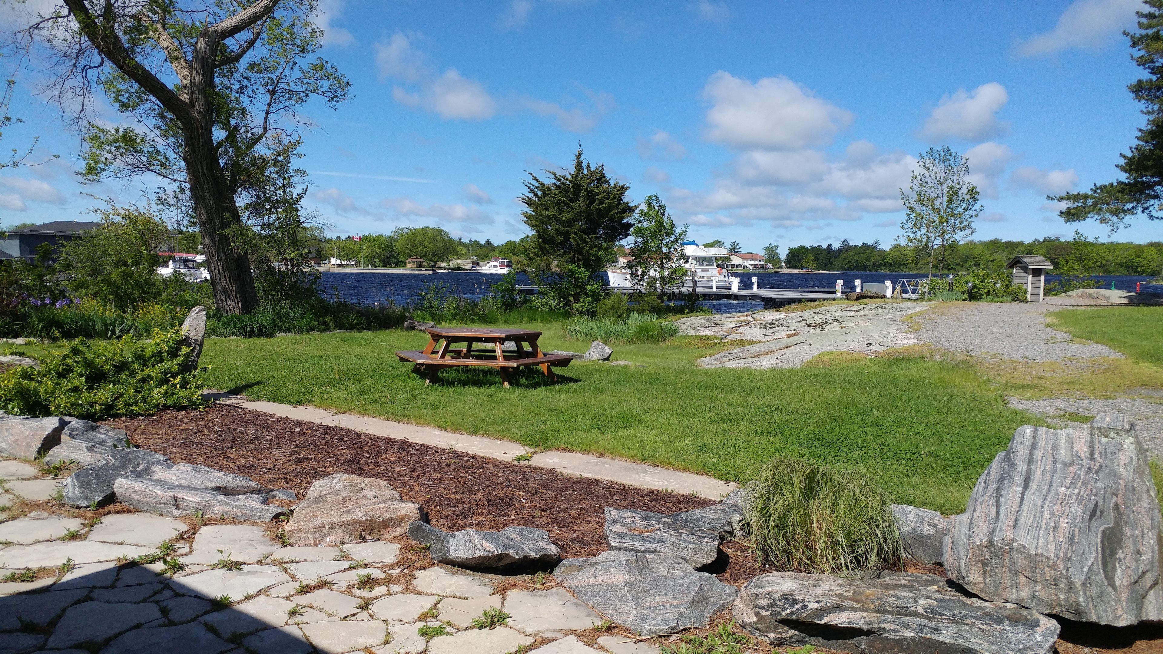 A picnic area with a table and benches, surrounded by rocks and grass, near a body of water with boats and buildings in the background.