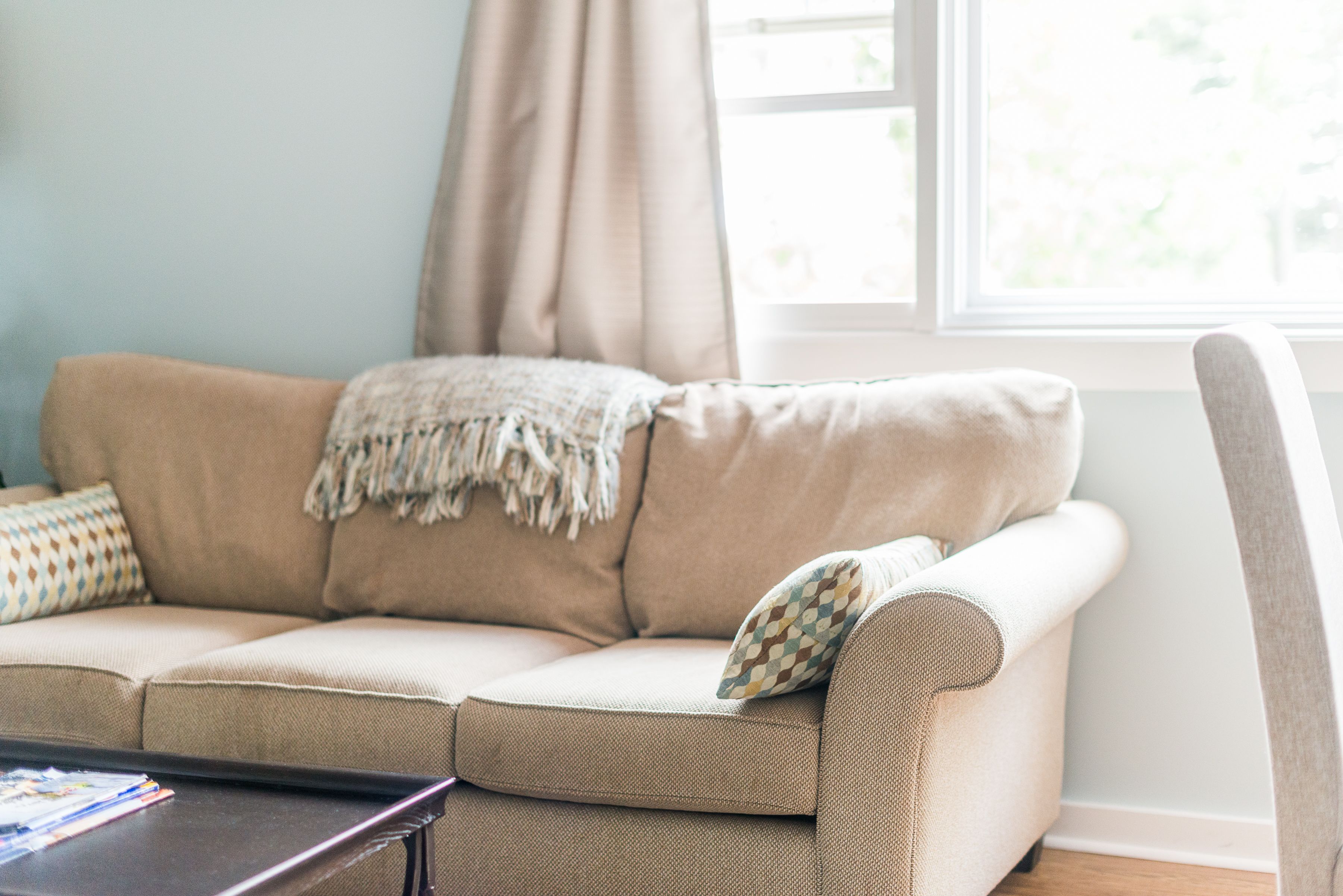 A beige sofa with a throw blanket and patterned pillows, positioned near a window with curtains.