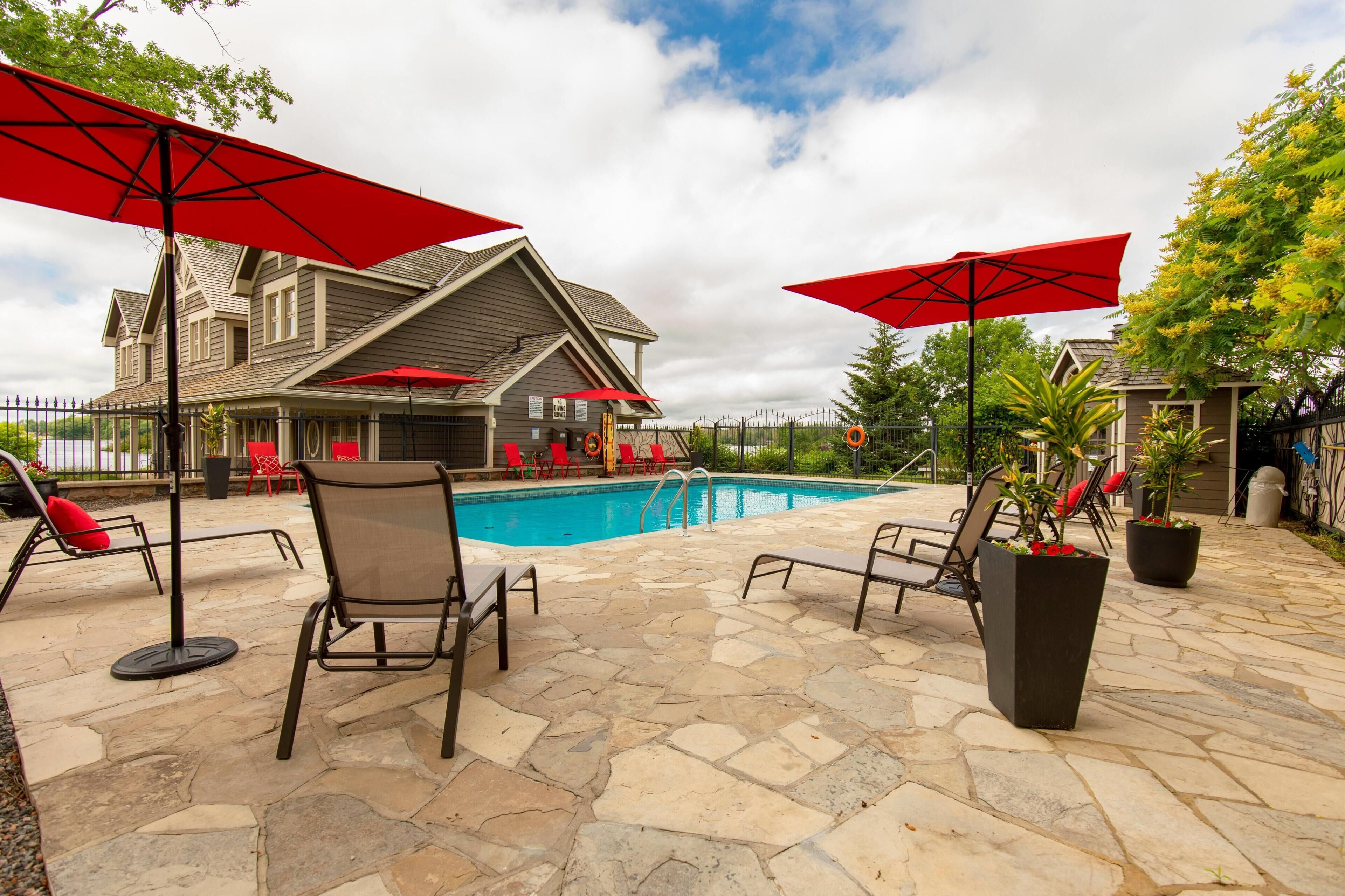 A stone patio with a pool, red umbrellas, and outdoor seating.