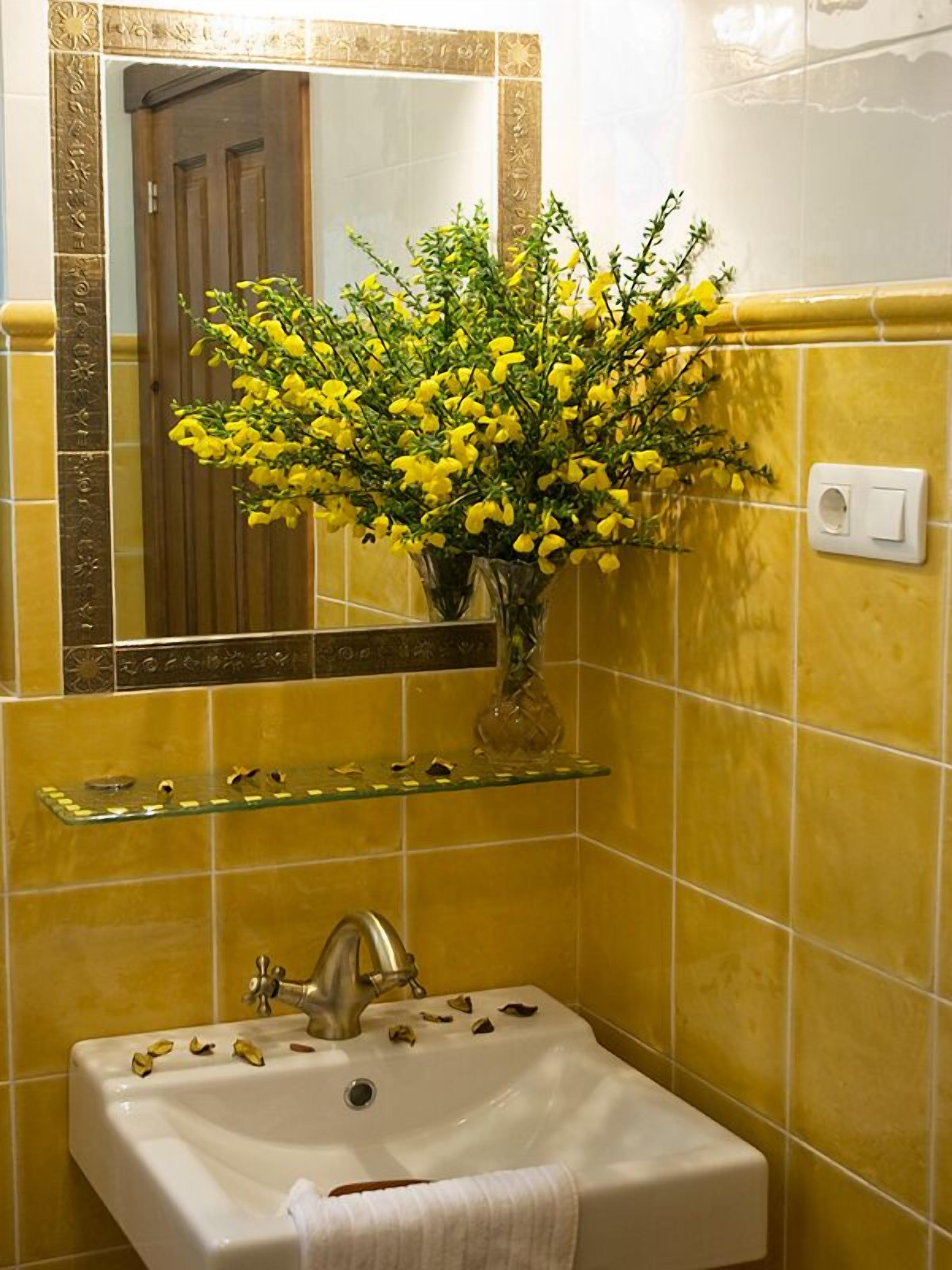 Um banheiro com paredes azulejadas em tons de amarelo, pia branca, prateleira de vidro com artigos de higiene pessoal e espelho com moldura decorativa.