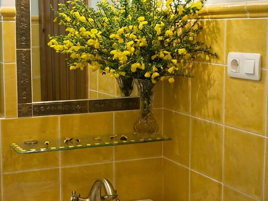 Um banheiro com paredes azulejadas em tons de amarelo, pia branca, prateleira de vidro com artigos de higiene pessoal e espelho com moldura decorativa.