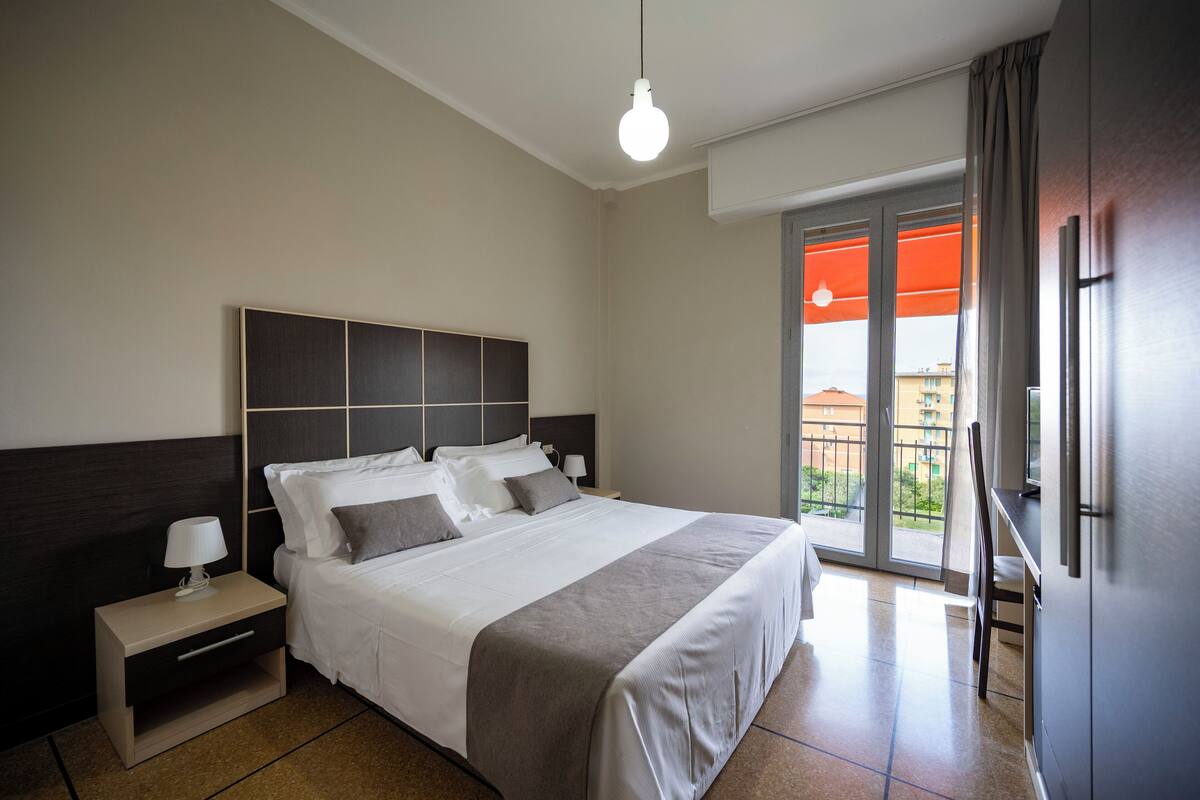 A modern hotel room with a large bed, a nightstand, a desk, and a balcony with an orange awning.