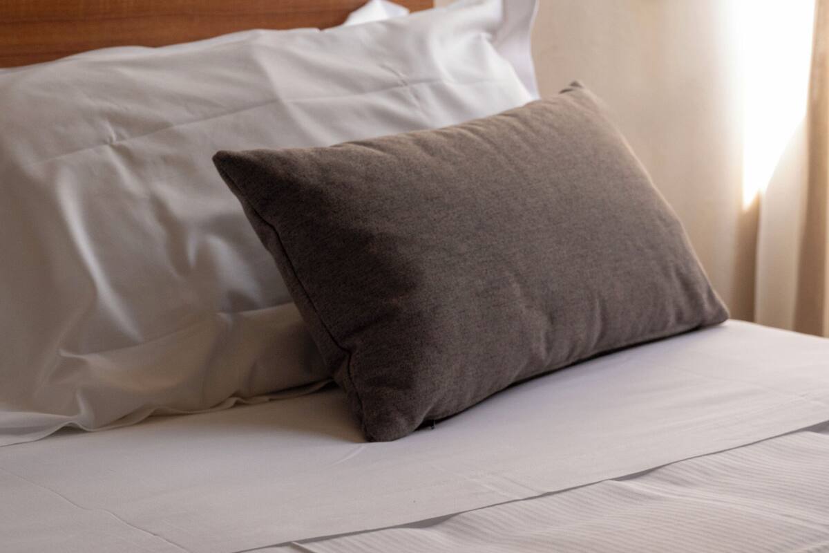 A neatly made bed with white and gray bedding, a wooden headboard, and a window with curtains.