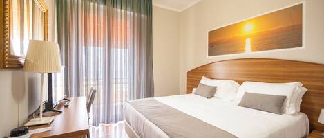 A hotel room with a large bed, a desk with a lamp, a chair, a desk lamp, a framed picture on the wall, and a window with curtains.