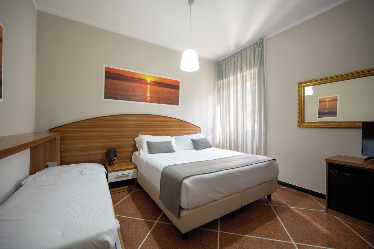 A hotel room with a bed, bedside table, lamp, and a painting of a sunset.