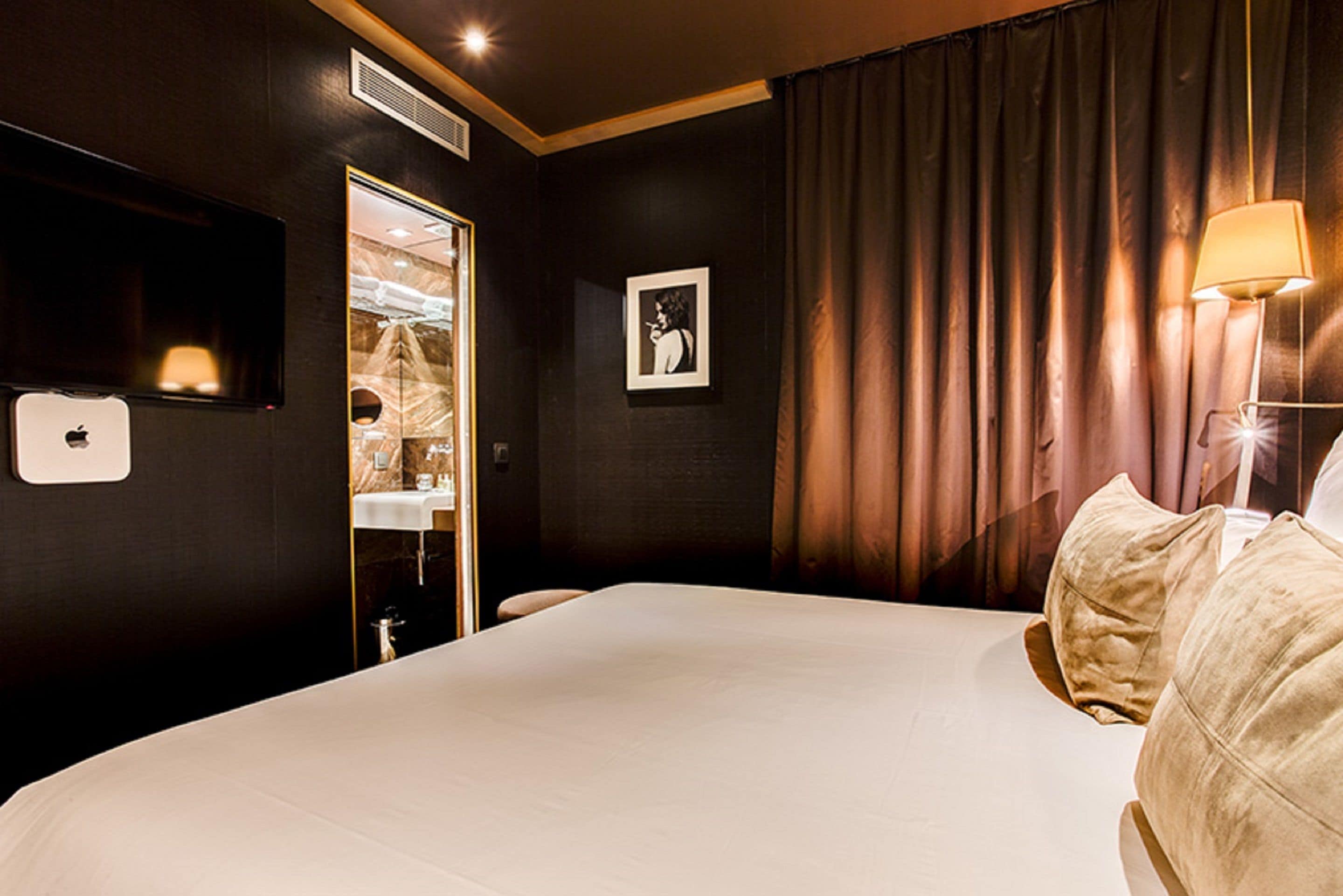 A hotel room with a large bed, a TV mounted on the wall, a mirror, a door leading to a bathroom, and a lamp on the bedside table.