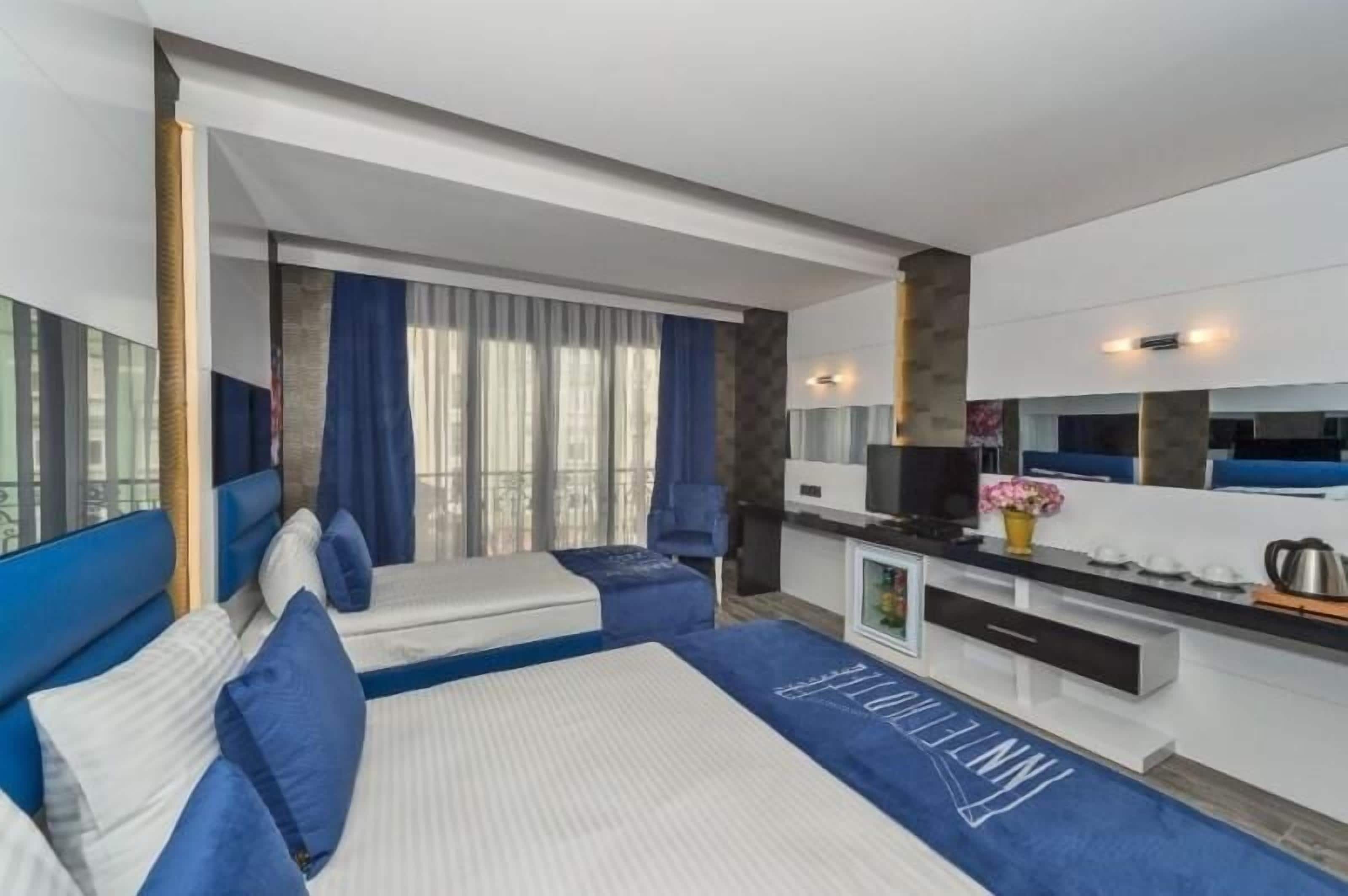 A modern hotel room with two beds, a kitchenette, and a large window with curtains.