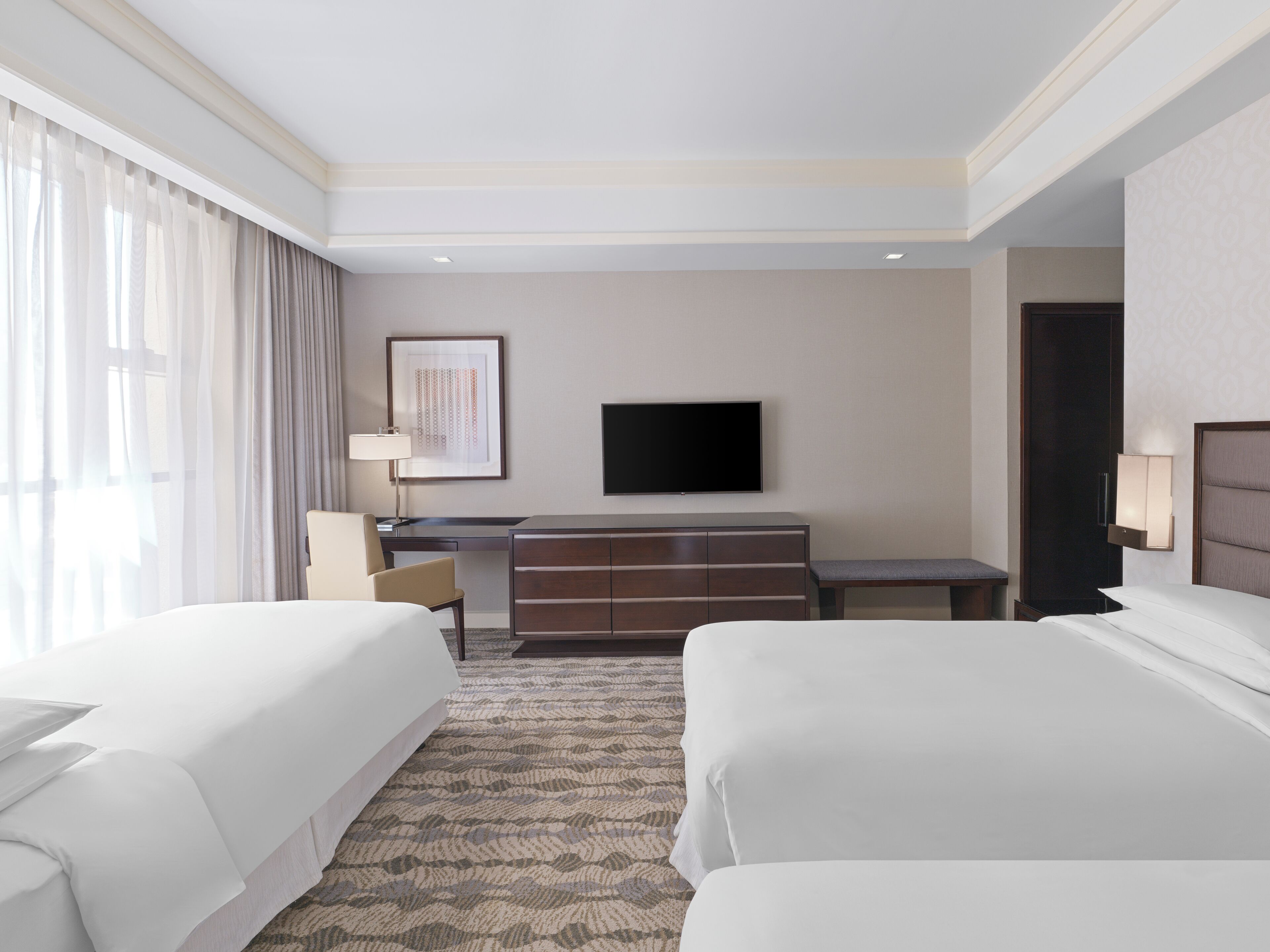 A hotel room with two beds, a desk, a chair, a television, and a large window with curtains.