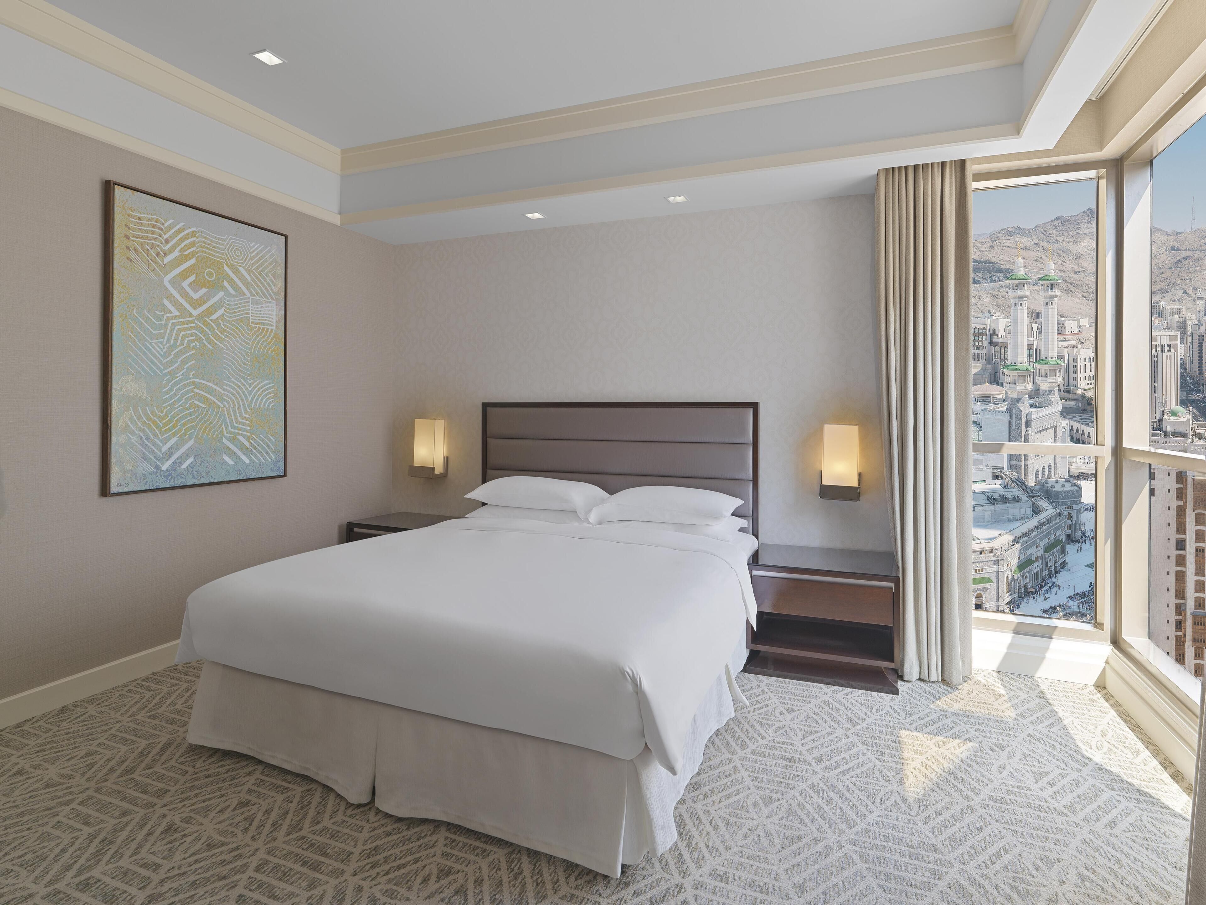 A hotel room with a large bed, two bedside lamps, a city view, and a patterned carpet.