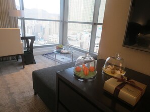 Ein Hotelzimmer mit Blick auf eine Stadtlandschaft durch große Fenster. Es gibt einen Schreibtisch mit einer Glaskuppel, die Orangen und eine Geschenkbox enthält.