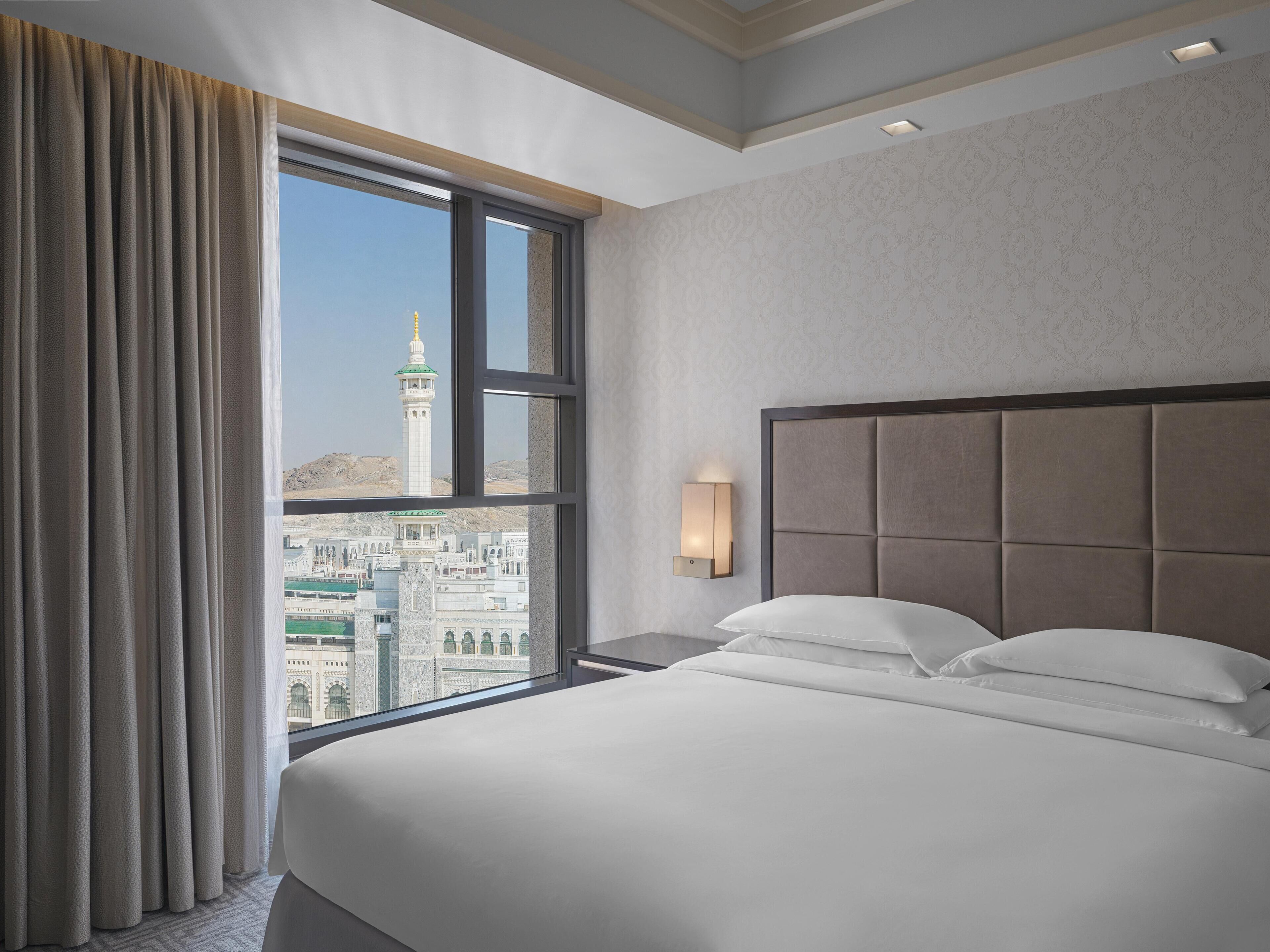 Ein Hotelzimmer mit einem großen Bett, einem Nachttisch, einer Wandlampe und Blick auf eine Stadtlandschaft durch ein Fenster.