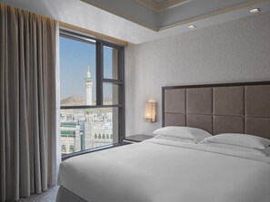 Habitación de hotel con una cama grande, una mesita de noche, una lámpara de pared y vistas a un paisaje urbano por la ventana.
