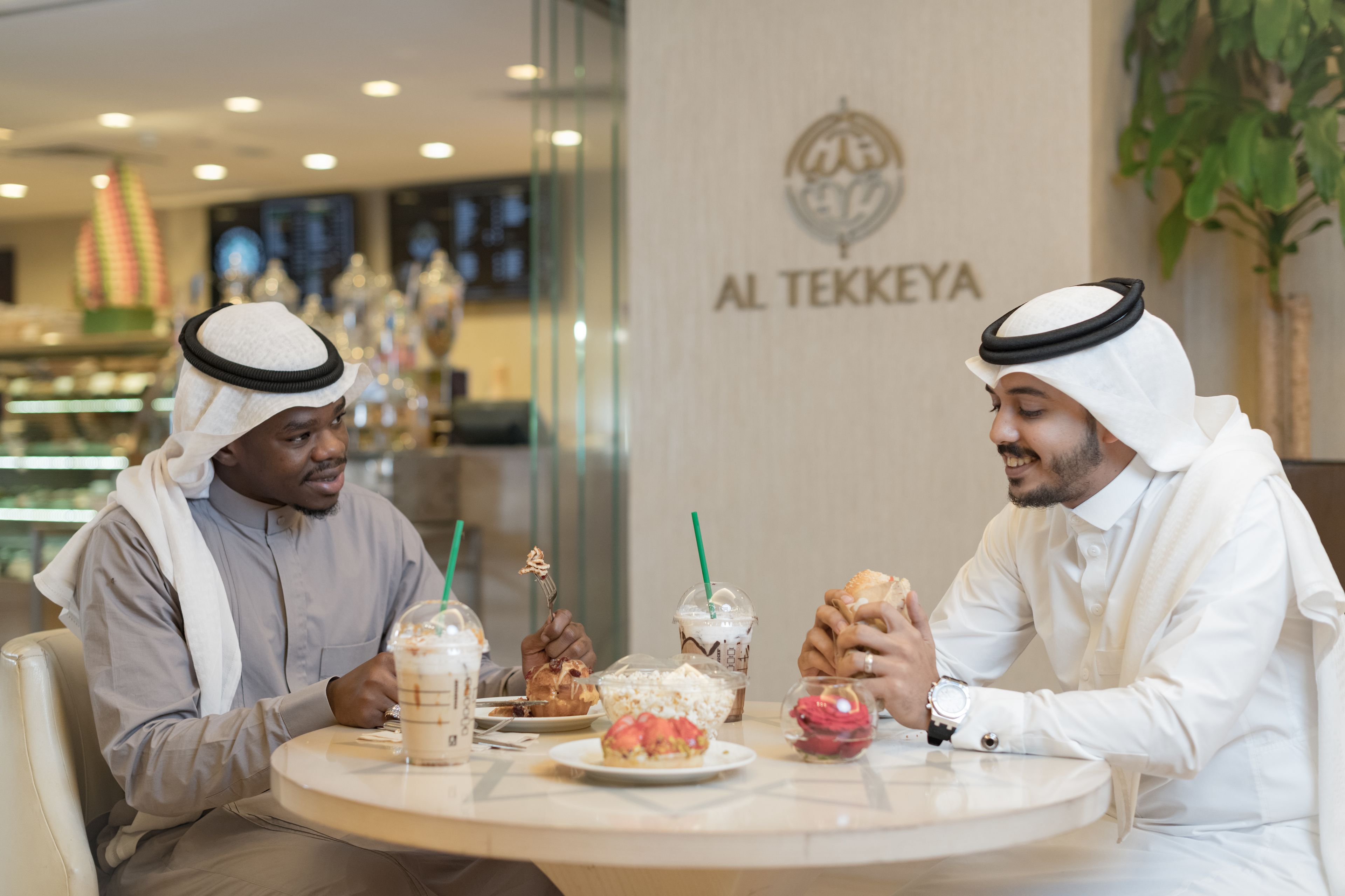 Two men dining at a table in a restaurant with a sign reading "AL TEKKEYA" in the background.