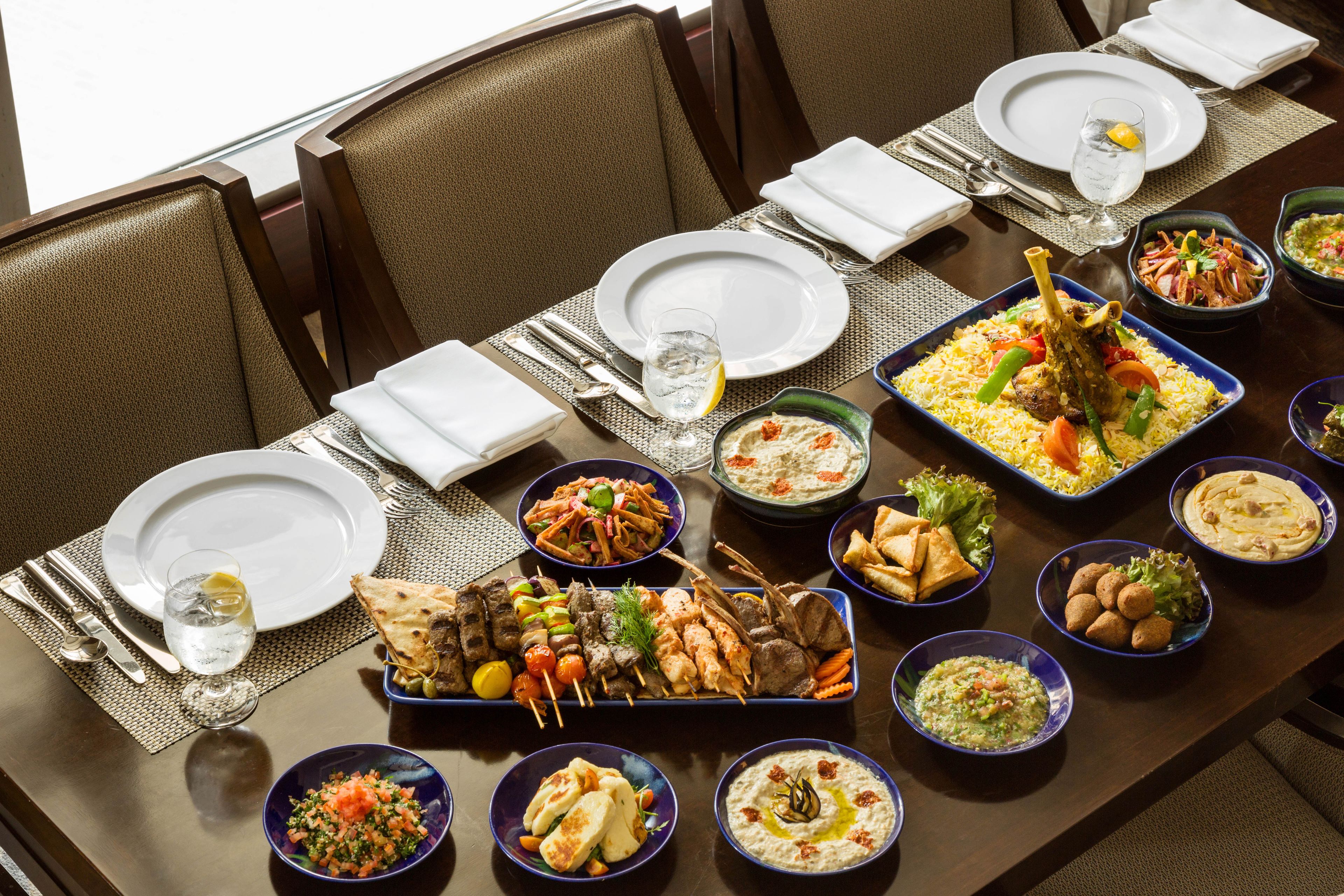 Una mesa de comedor dispuesta con diversos platos, incluyendo una bandeja grande de carnes y verduras, tazones de hummus y un vaso de agua.