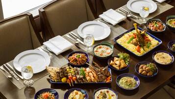 Una mesa de comedor dispuesta con diversos platos, incluyendo una bandeja grande de carnes y verduras, tazones de hummus y un vaso de agua.
