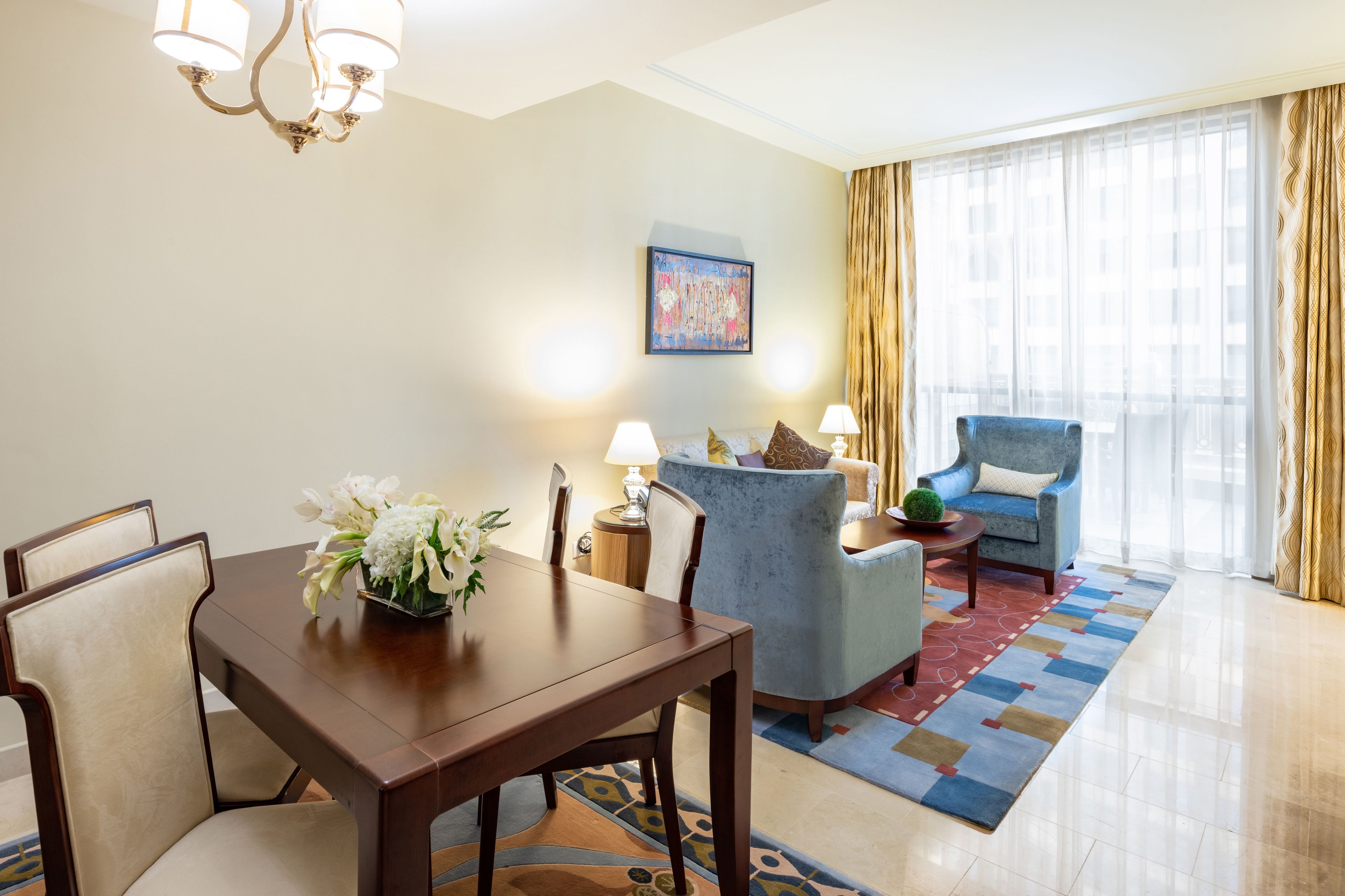 A hotel room with a dining table, chairs, a blue armchair, a small side table with a lamp, and a large window with curtains.