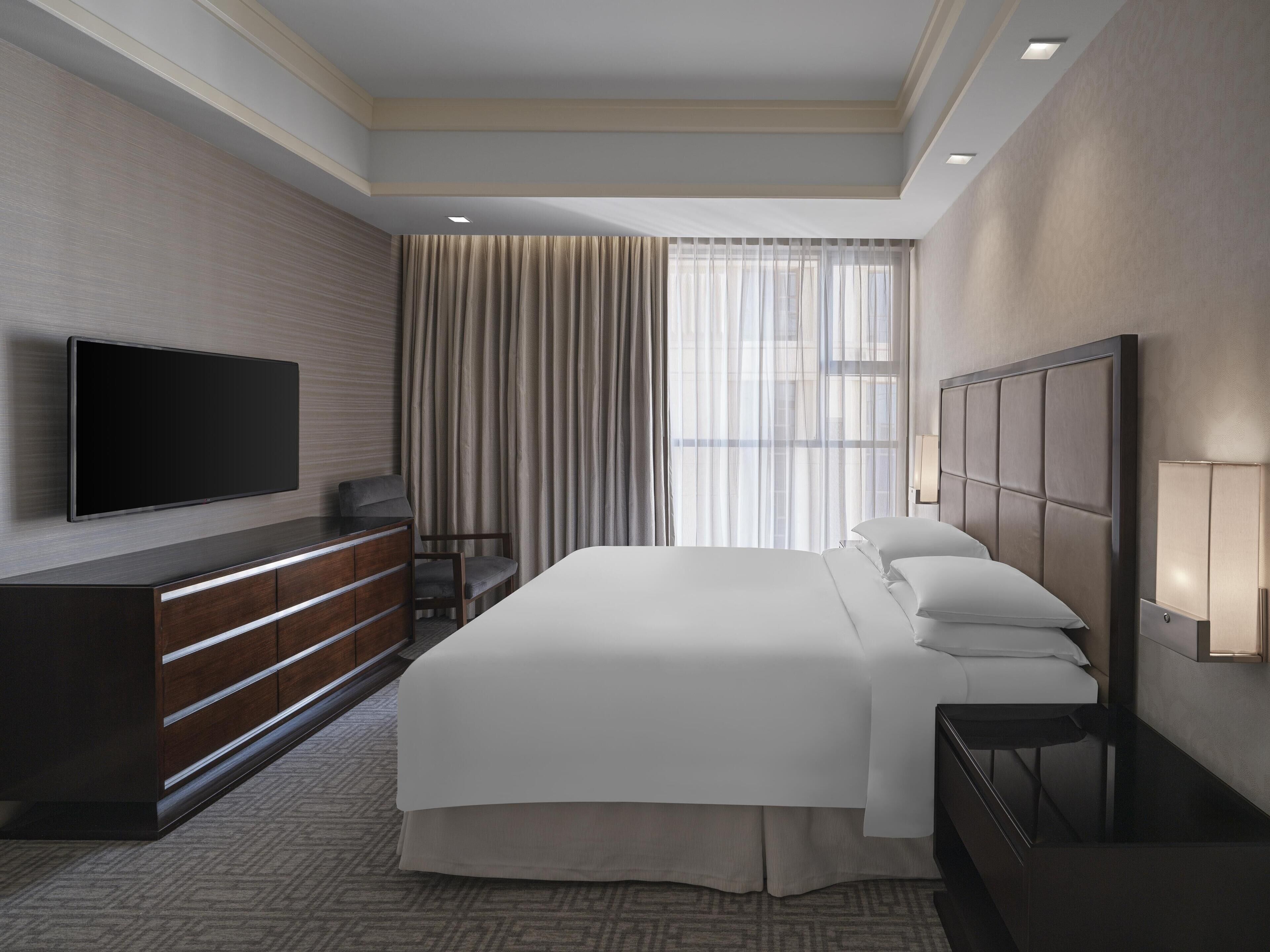 A hotel room with a large bed, a flat-screen TV mounted on the wall, a bedside table, and a window with curtains.