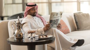 A man in traditional attire reading a newspaper in a well-lit room with a sofa and a small table with a teapot and snacks. - Jabal Omar Hyatt Regency Makkah (Makkah)
