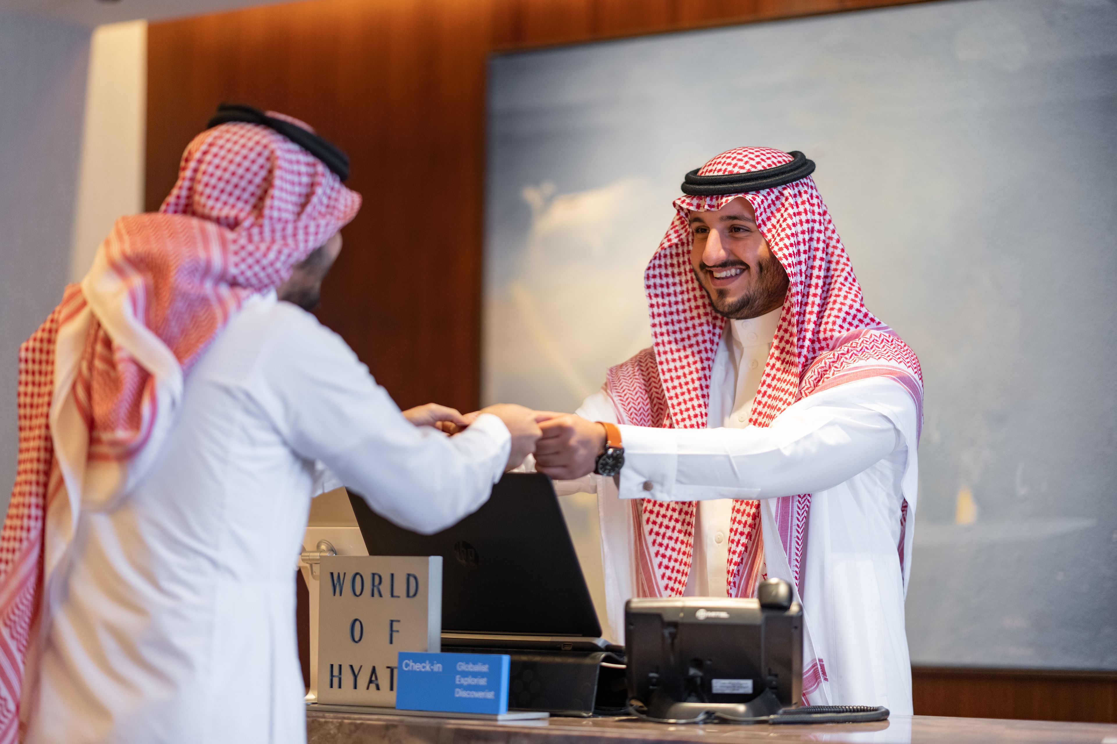 Dos hombres estrechan la mano en el vestíbulo de un hotel, con un cartel que dice "WORLD OF HYAT" y una computadora portátil sobre el mostrador.