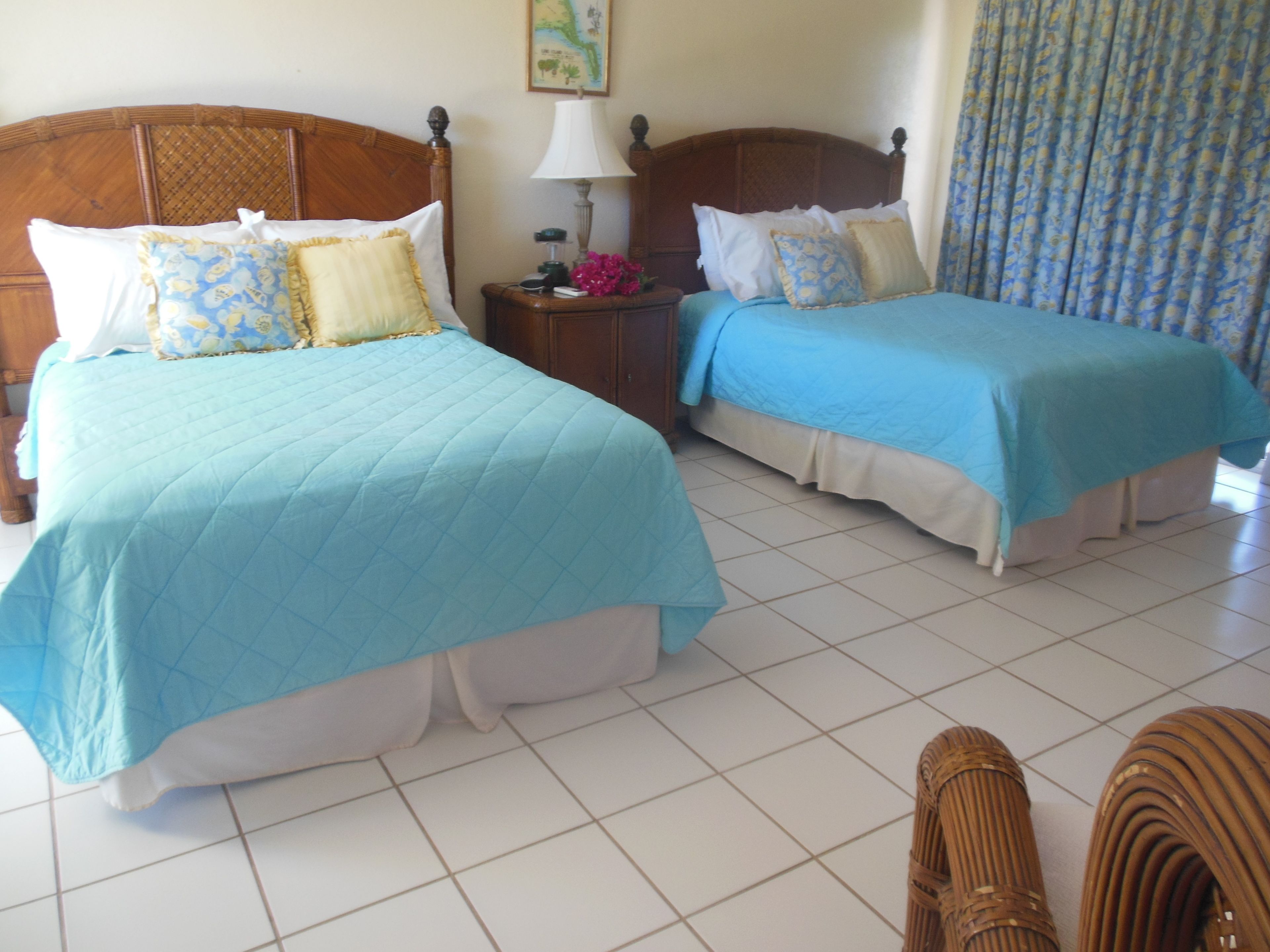 Habitación de hotel con dos camas, una mesita de noche con lámpara y una silla de mimbre.