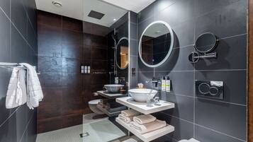 Un baño moderno con azulejos oscuros, un box de ducha de vidrio, un lavabo blanco y dos espejos fijos en la pared.