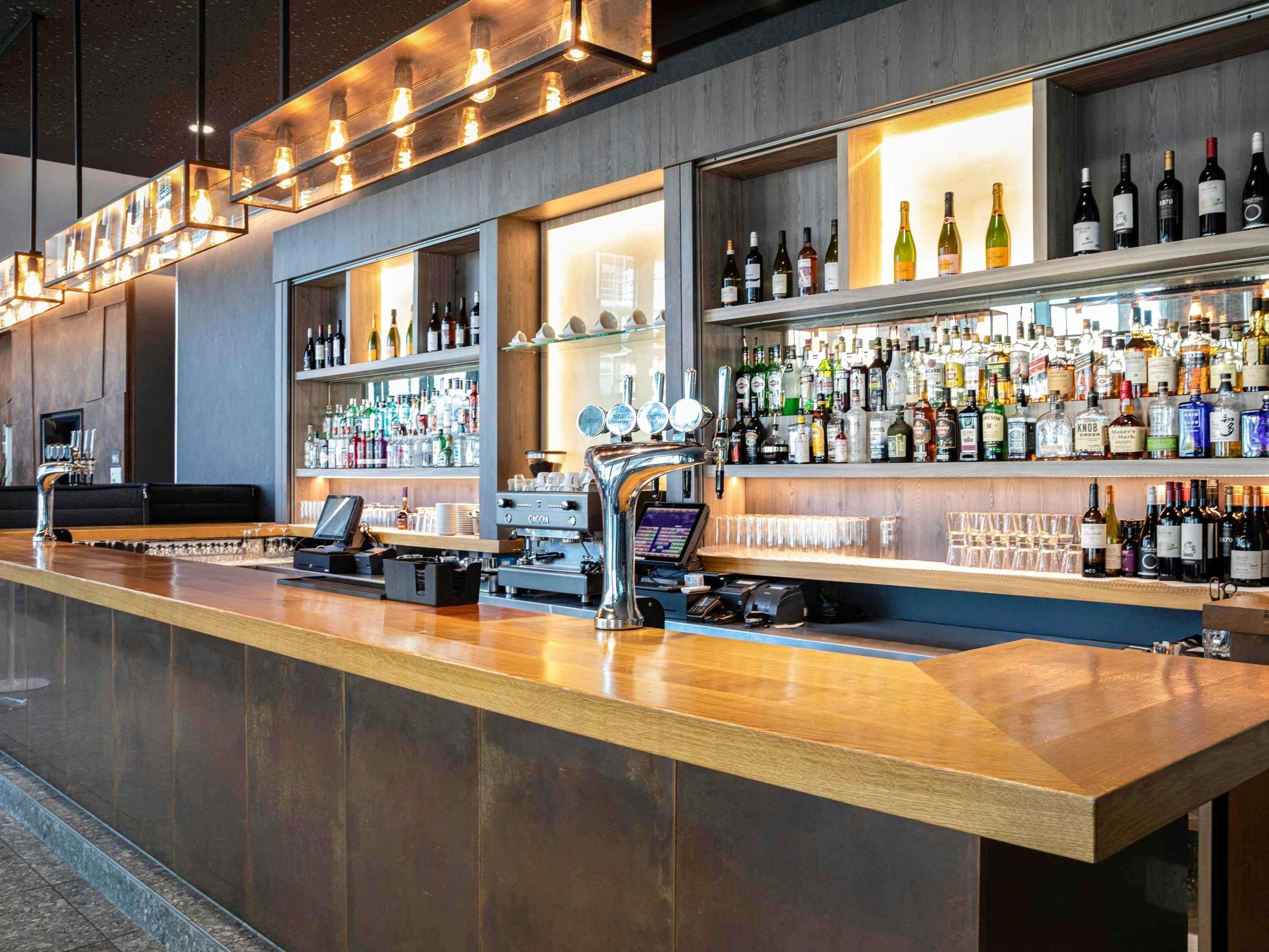 Uma área de bar com balcão, banquetas, um registrador de caixa e diversas garrafas de bebidas alcoólicas.