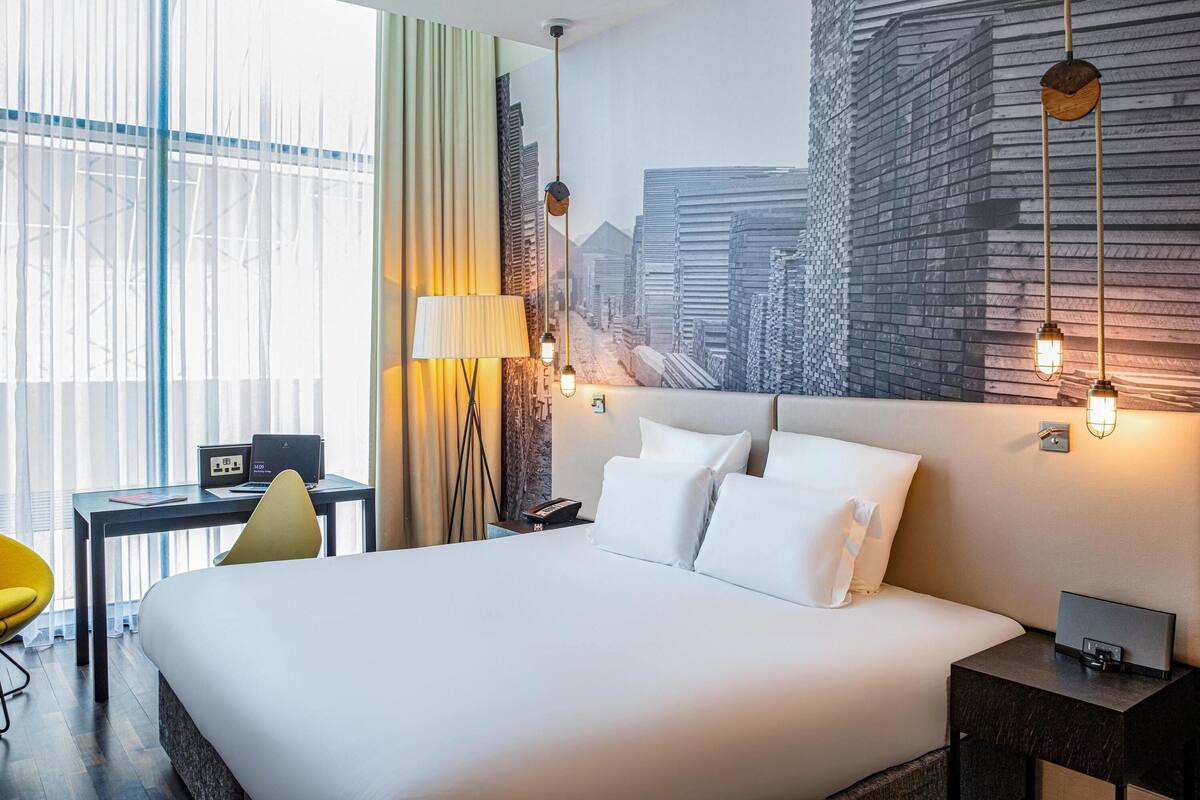 Une chambre d’hôtel moderne avec un grand lit, un bureau avec un ordinateur portable, une chaise et une fresque représentant un paysage urbain sur le mur.