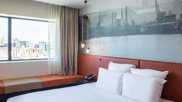 A hotel room with a large bed, a view of a cityscape through a window, a wall-mounted telephone, and a seating area with a view.